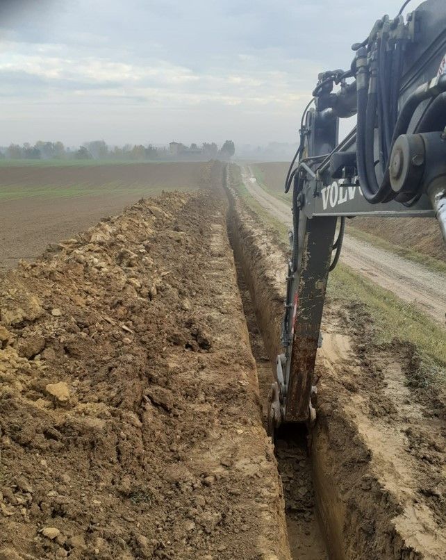 Un escavatore scava una trincea lungo una strada sterrata in un campo rurale sotto un cielo coperto.