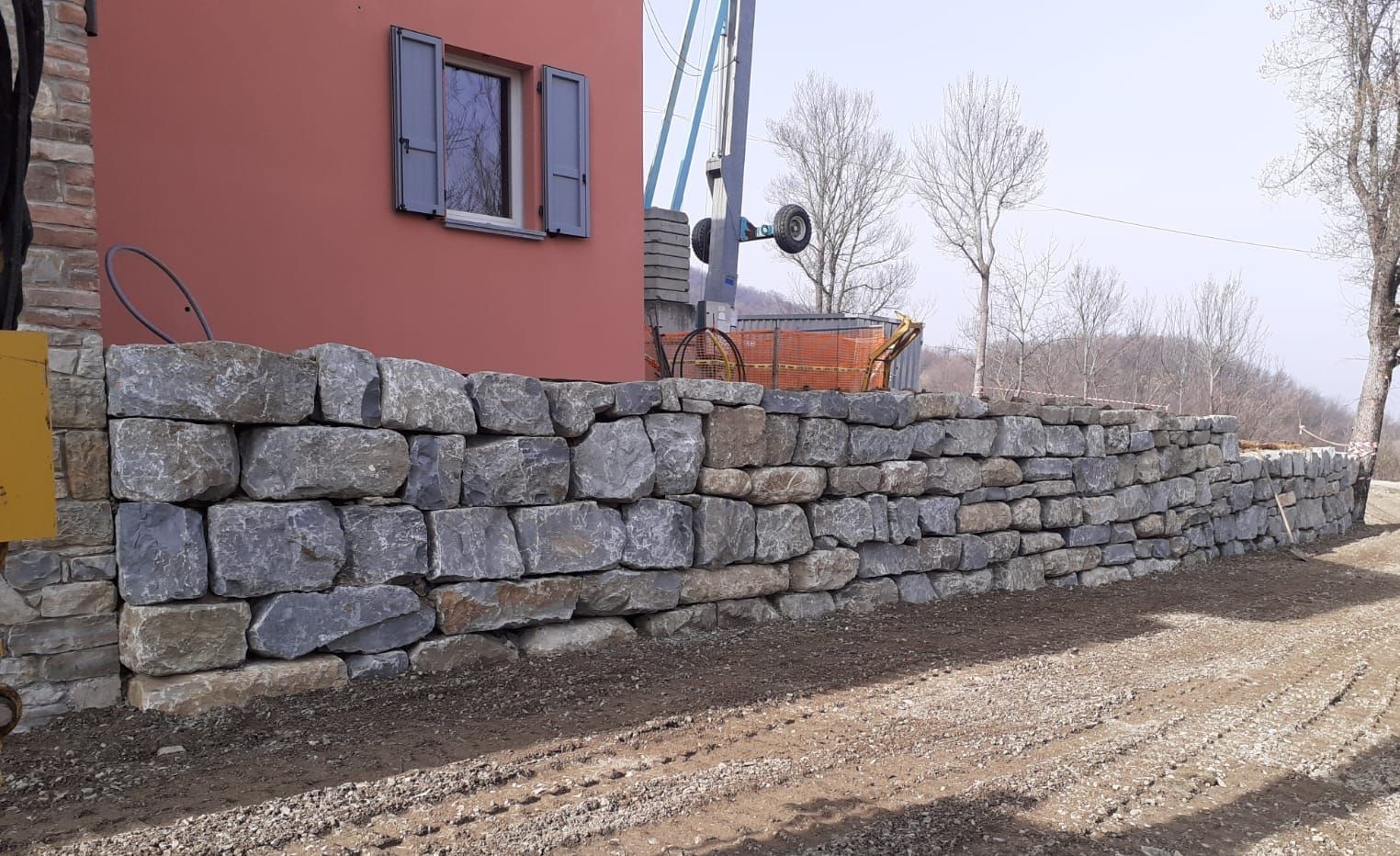 Muro di pietra di fronte a un edificio rosso con una piccola finestra e persiane blu. Strada sterrata in primo piano.