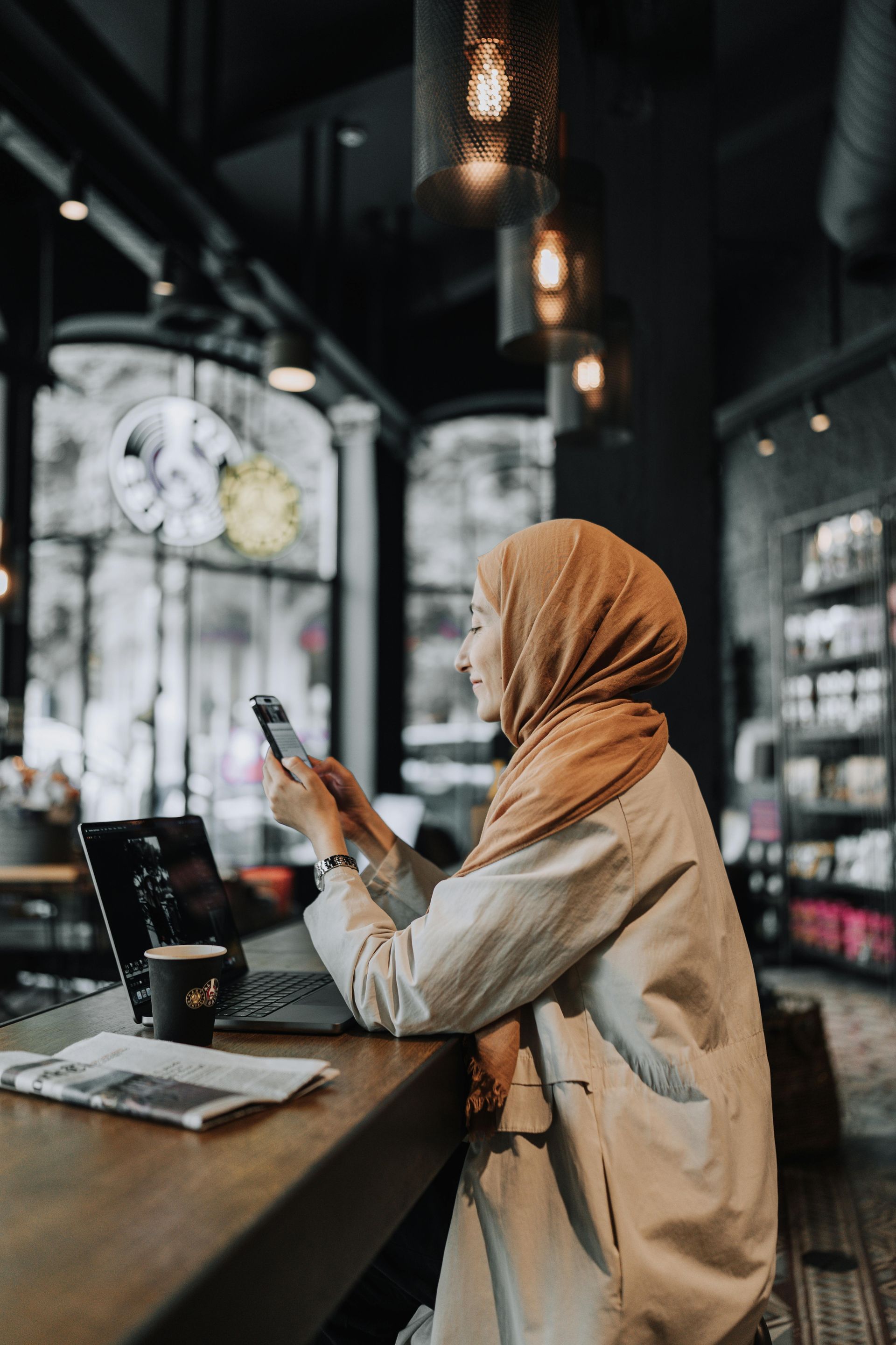 A woman in a hijab is sitting at a table in a cafe looking at her phone.