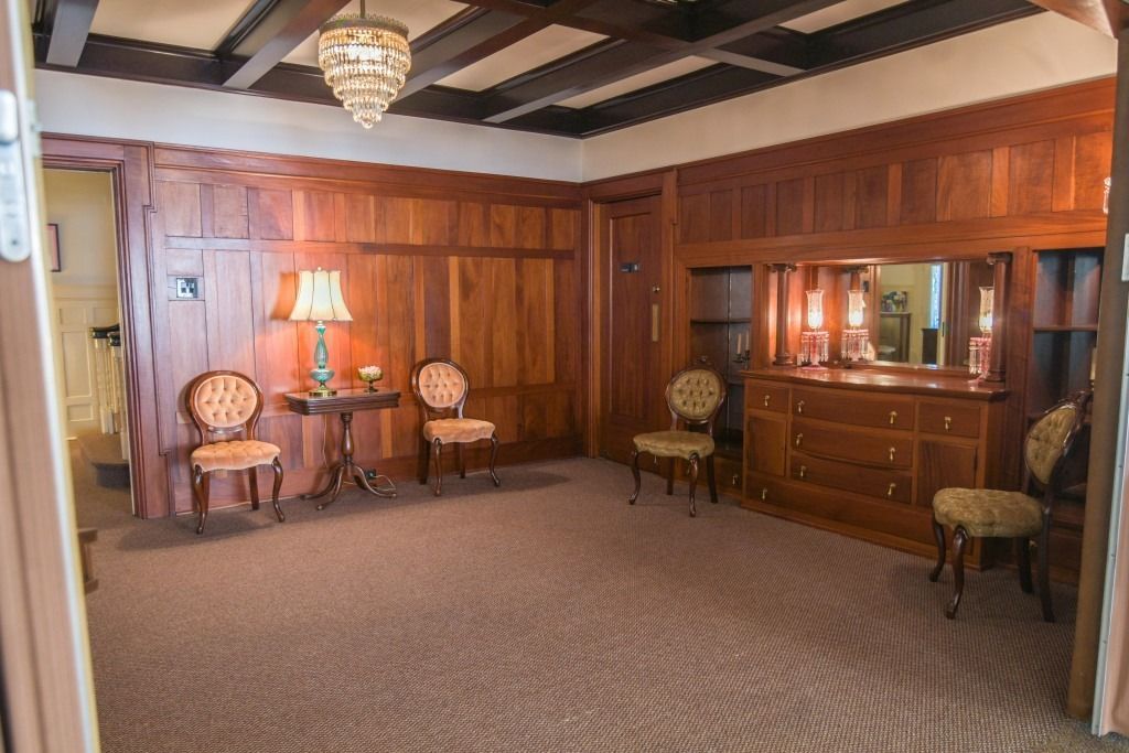 Empty vintage room with wood-paneled walls, patterned carpet, chandelier, and antique chairs around a small table