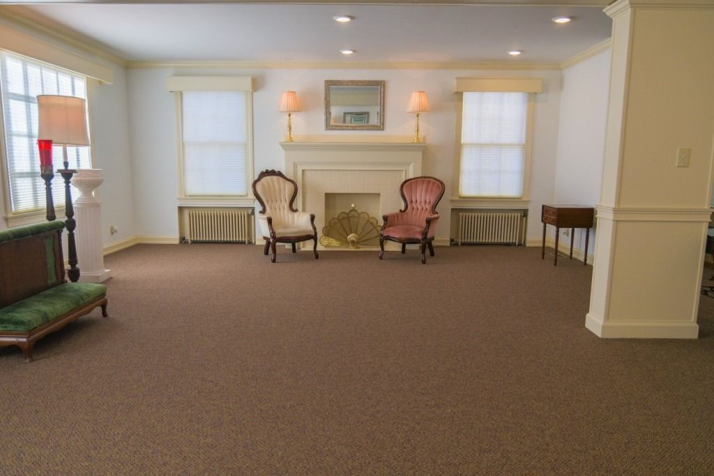 Empty carpeted room with two armchairs by a fireplace and tall windows, softly lit by wall lamps