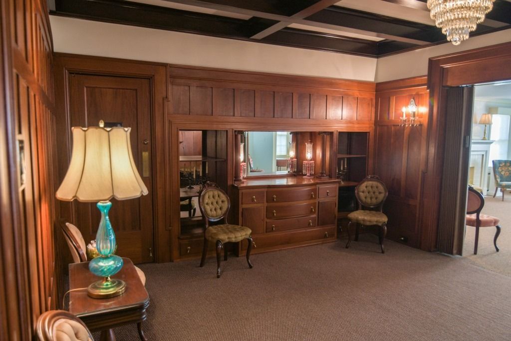 Wood-paneled room with chandeliers, antique chairs, a lamp, and a mirrored wall cabinet