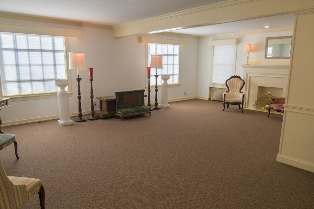 Empty living room with beige carpet, fireplace, large windows, and a few lamps and chairs.