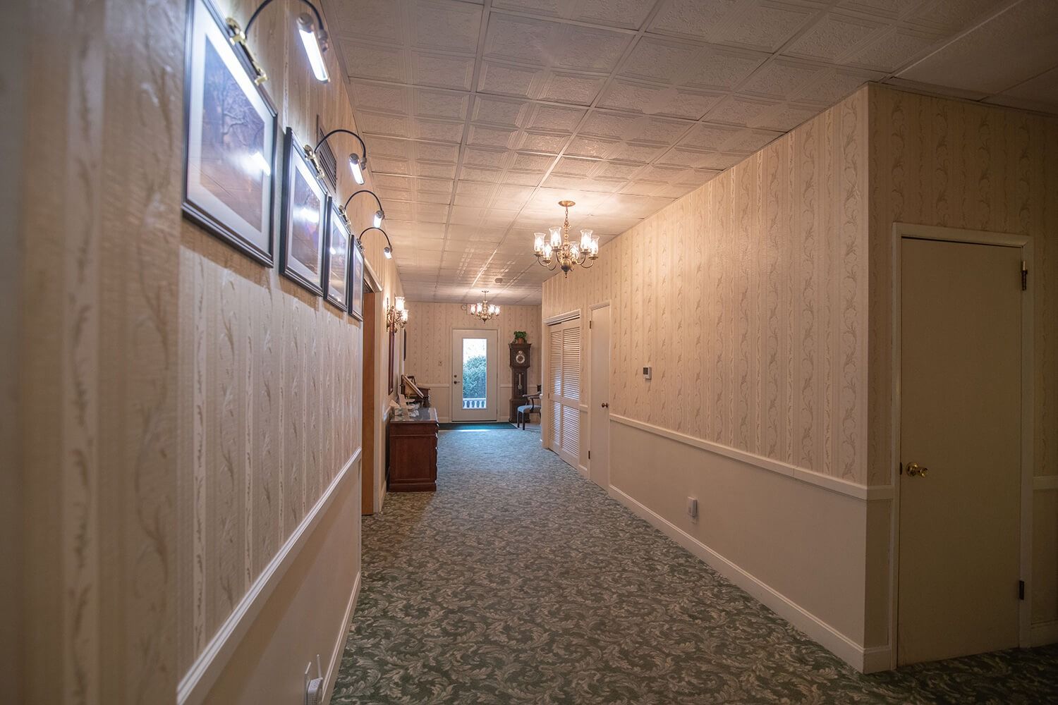 Long carpeted hallway with framed pictures, beige walls, and ceiling lights leading to a door.