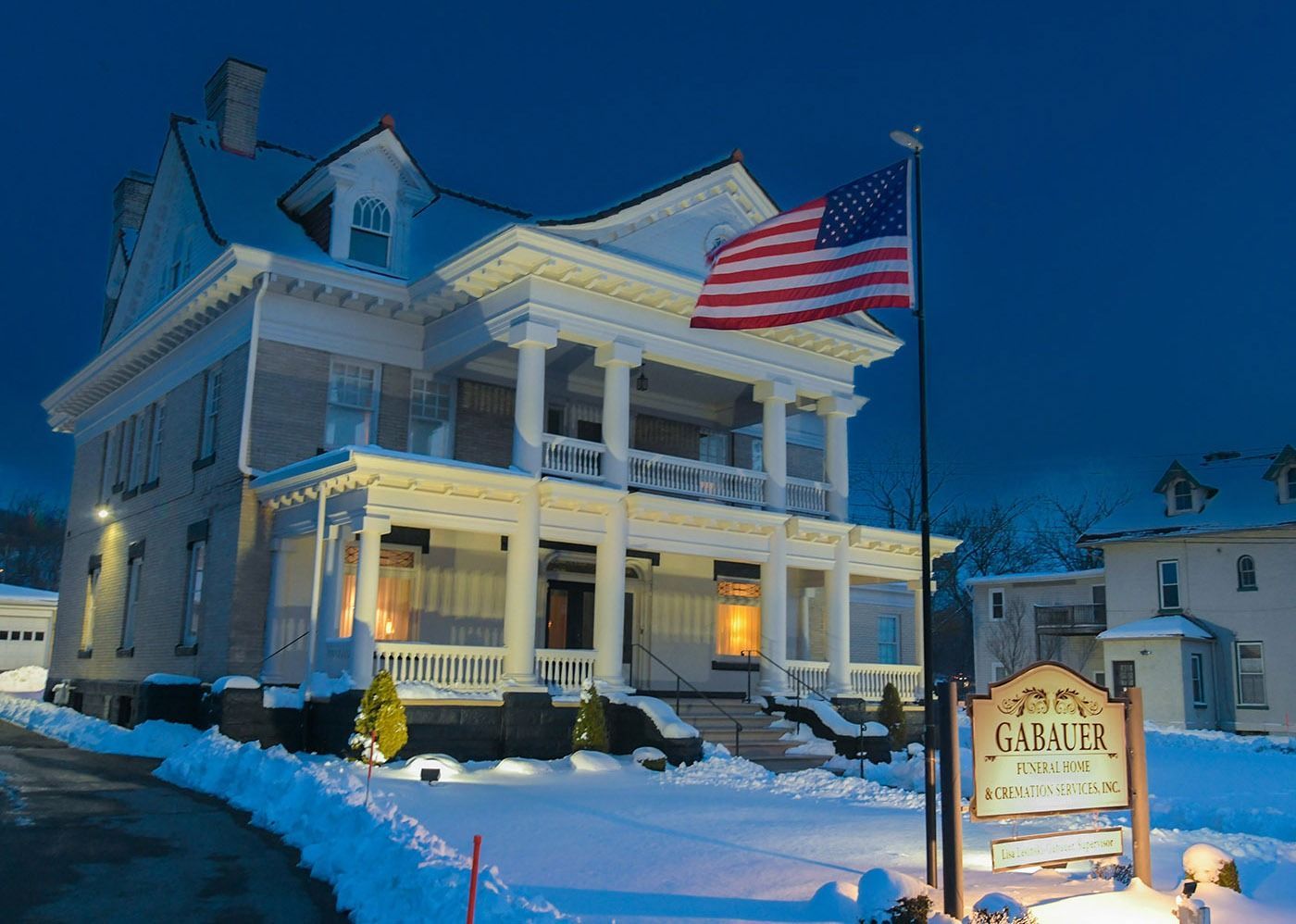 Snowy white inn at dusk with lit windows and a large American flag flying outside