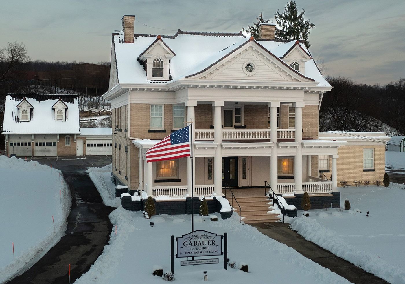 Snow-covered bed and breakfast with an American flag and lit porch at dusk