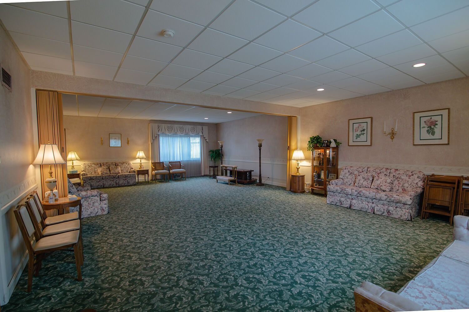 Spacious lounge with floral couches, chairs, lamps, and patterned carpet under a low white ceiling.