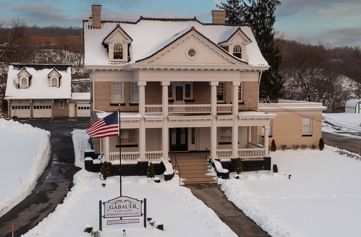 Snow-covered historic house with a porch and American flag in front of a driveway