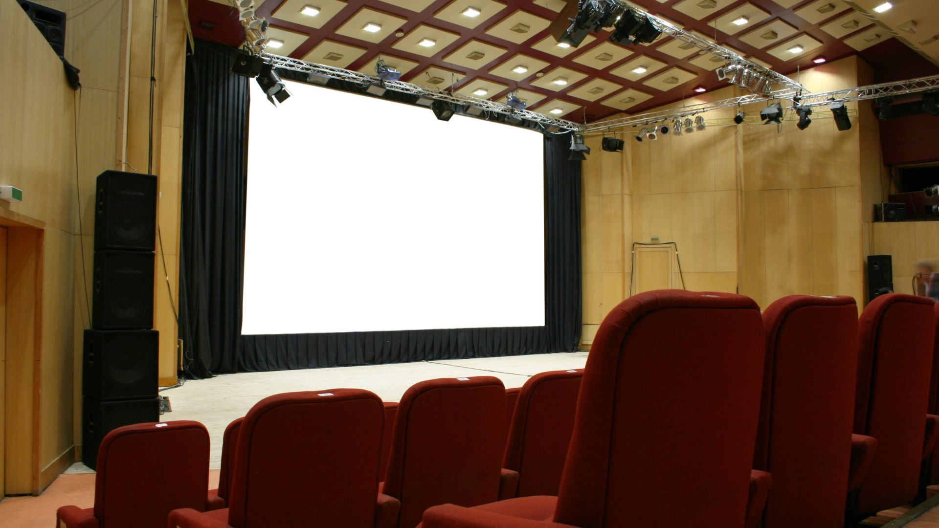 Empty auditorium with red seats facing a large white projection screen and stage curtains.