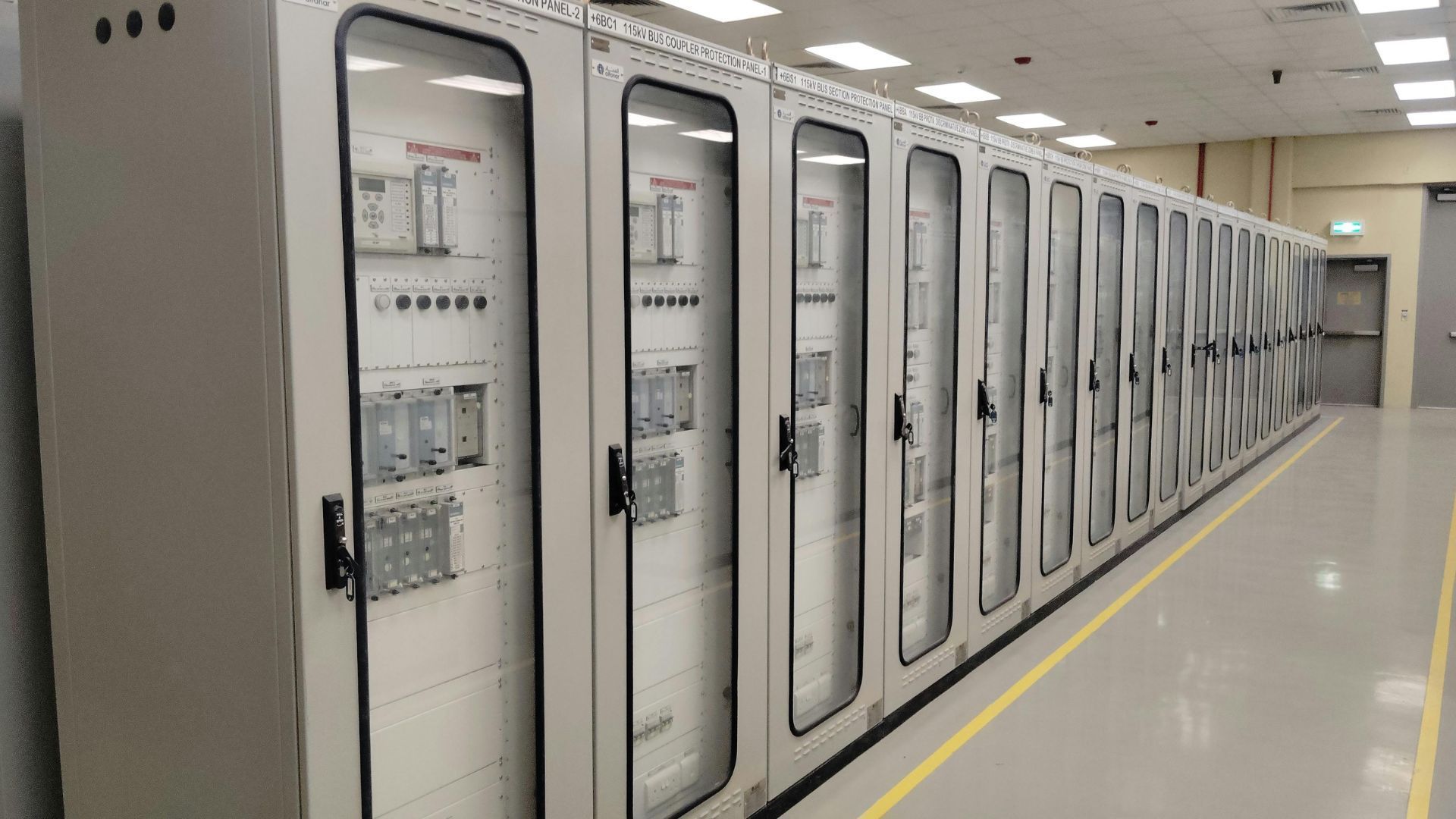Row of electrical control panels in a bright industrial corridor, with a yellow floor line along the wall.