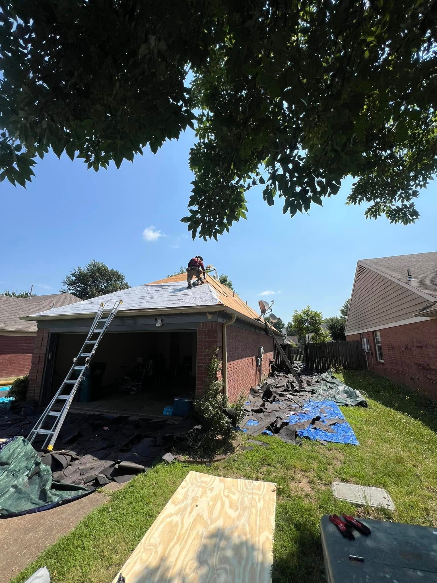 Workers repairing roof on a brick house, with ladders, tarp, and debris in the yard.