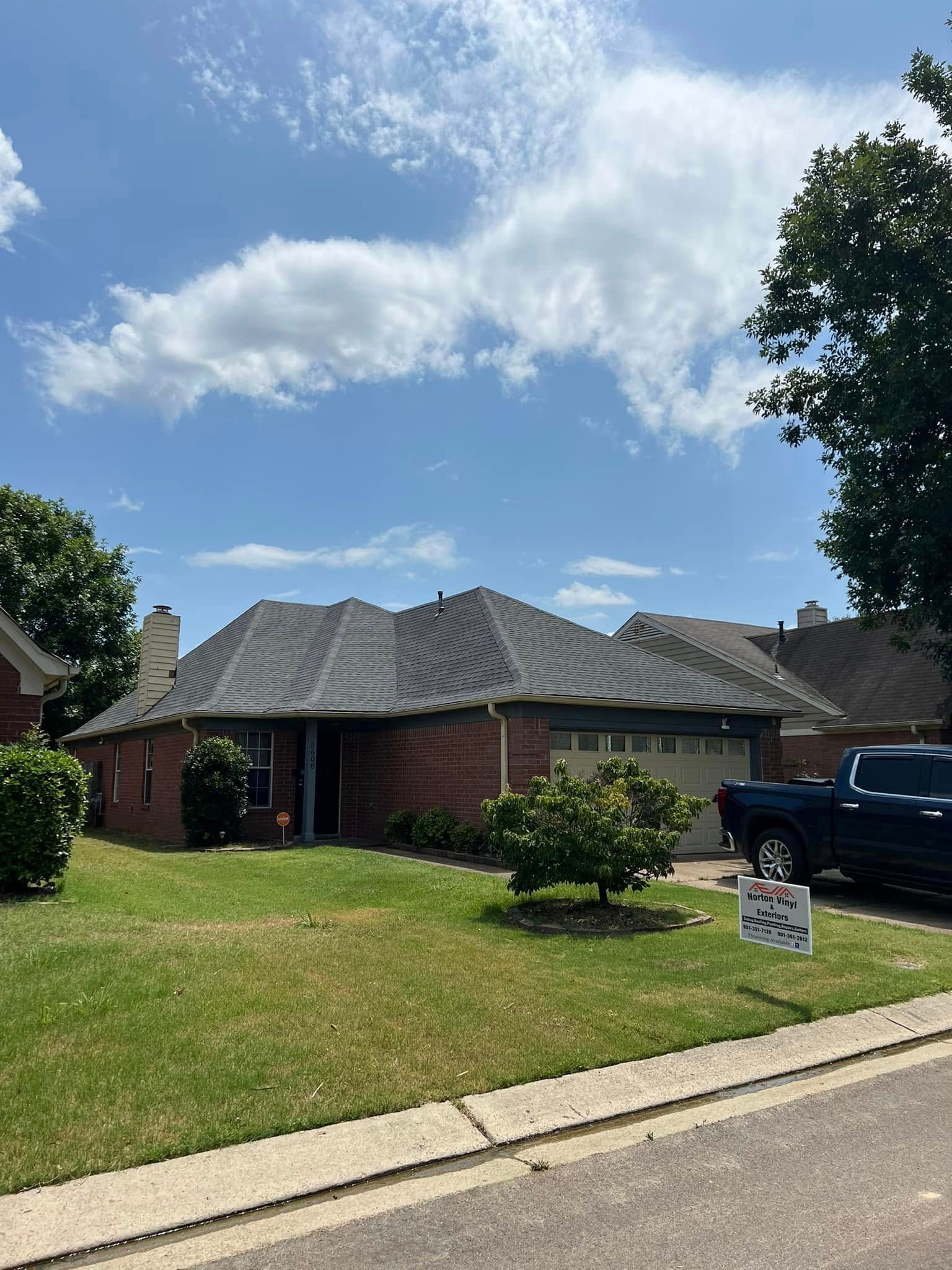 Suburban brick house with a dark roof, green lawn, blue sky, and a parked black truck by the curb