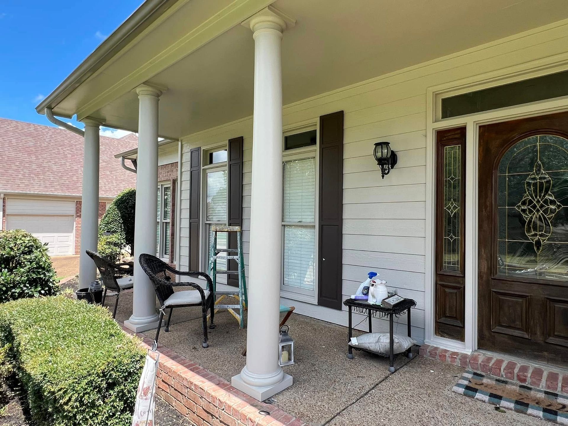 Front porch with white columns, wooden front door, small table, chairs, and neatly trimmed hedges