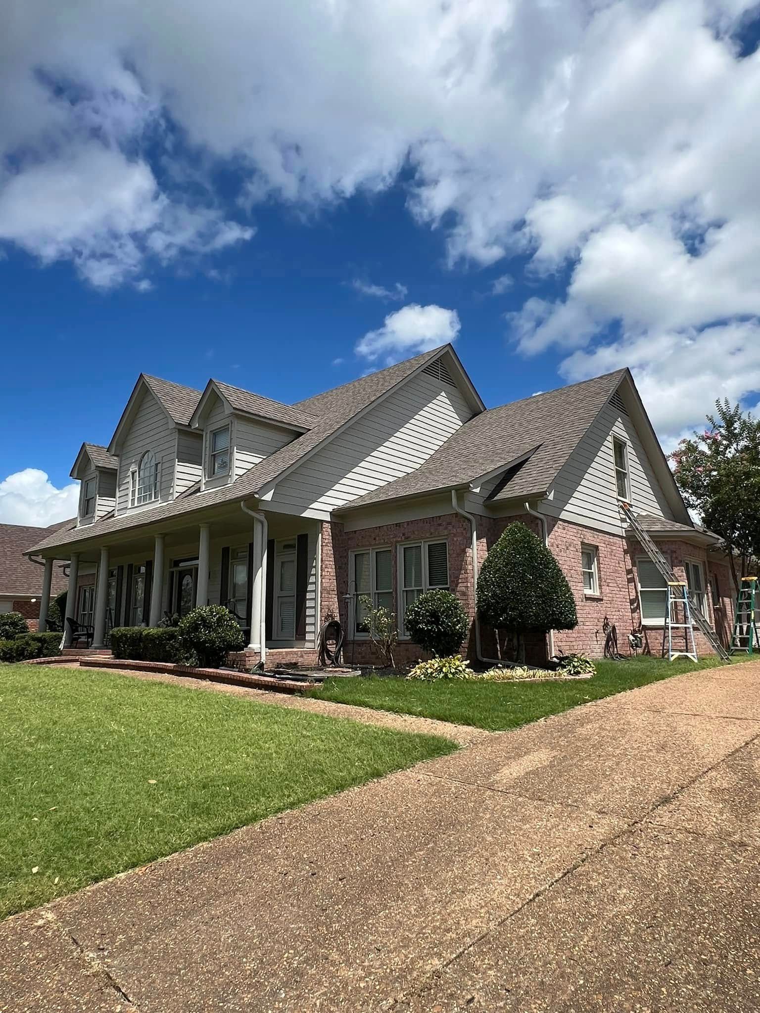 Large brick house with gabled roof, white porch columns, and a gravel driveway under a partly cloudy blue sky