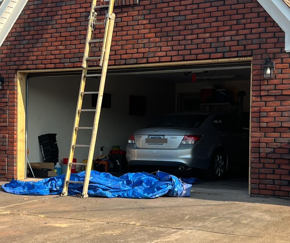 Garage with a silver car inside, a ladder leaning against the roof, and a blue tarp on the driveway.