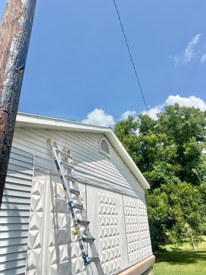 Ladder leaning against a white house under a blue sky, with a utility pole and trees nearby
