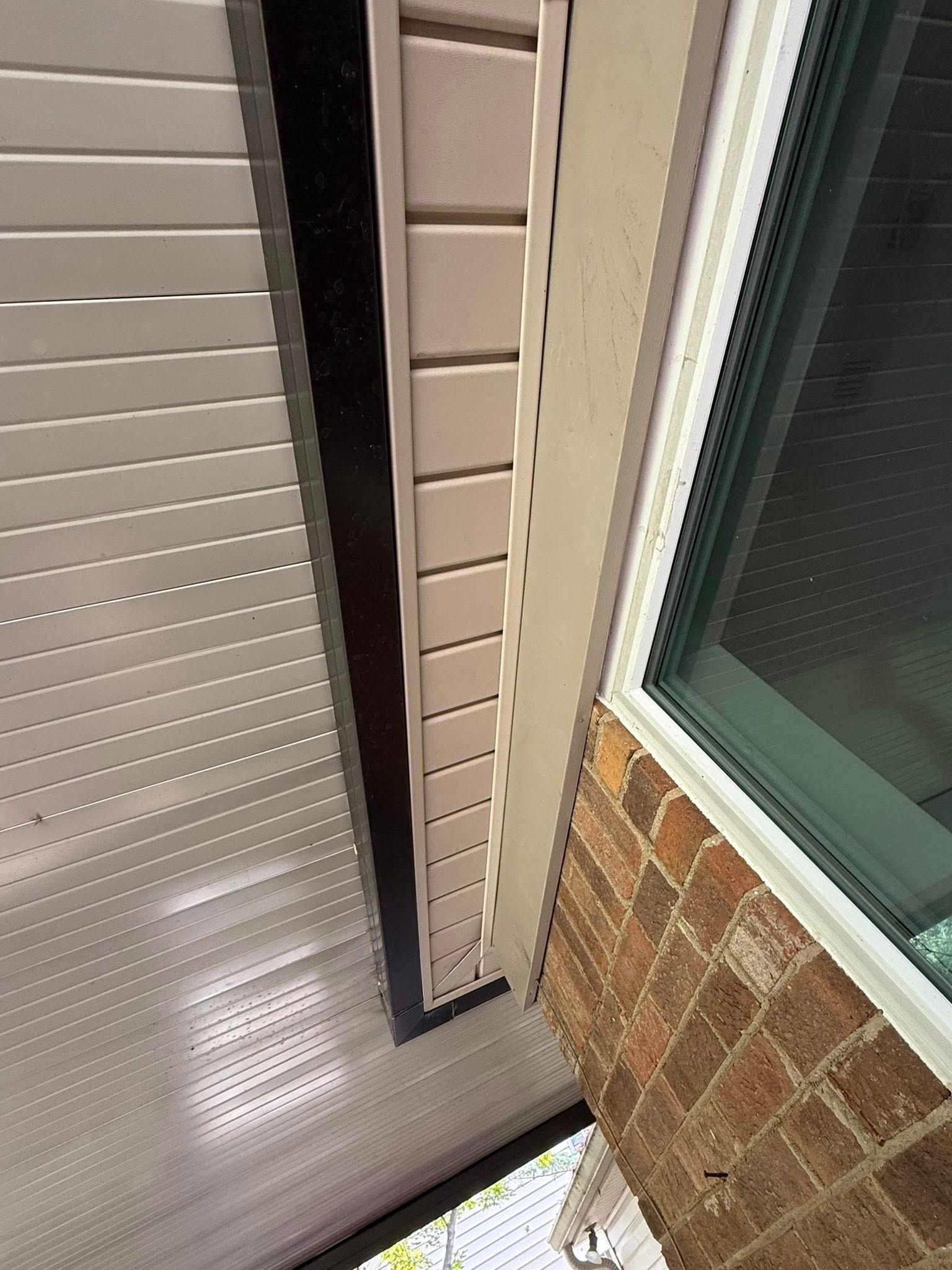 Close-up of a narrow gap between a metal awning, vinyl siding, and a brick wall beside a window.