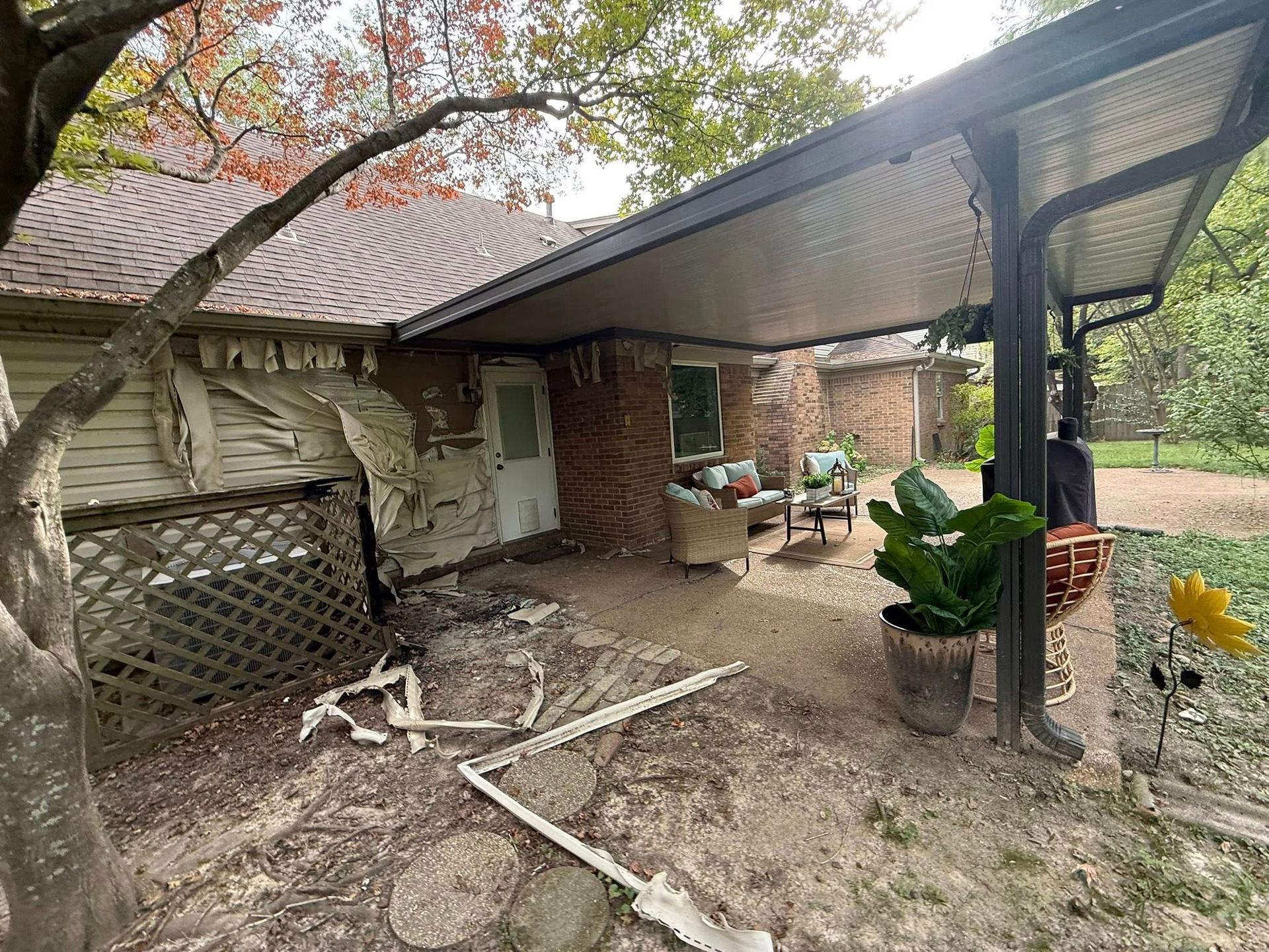 Backyard patio with brick house, pergola, potted plants, and scattered debris on dirt ground.