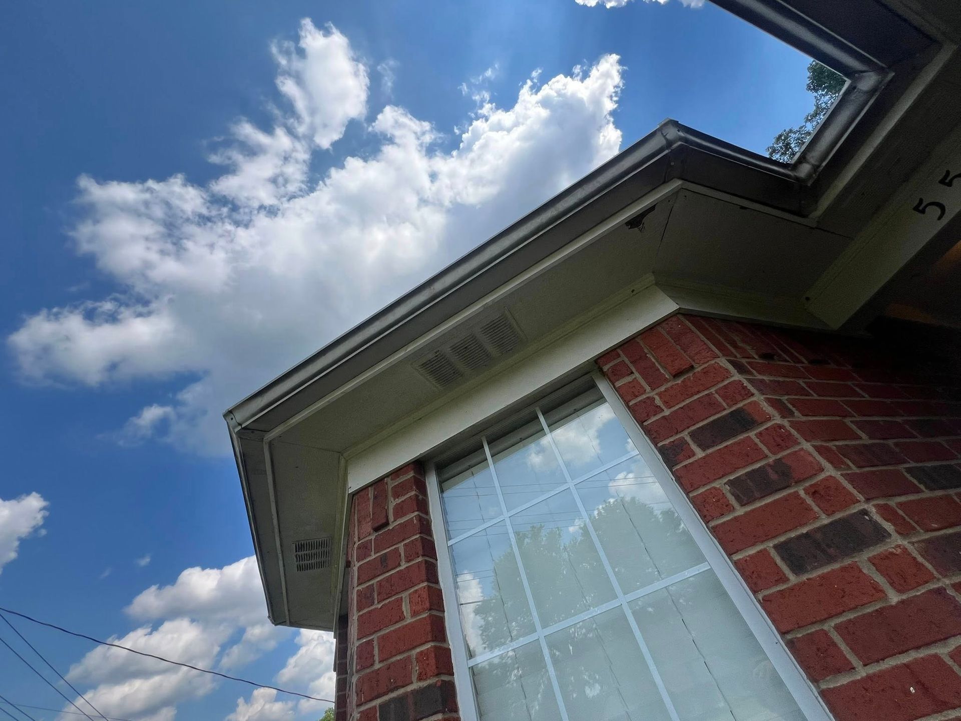 Brick house corner with white-trimmed window and roof against a blue sky with clouds