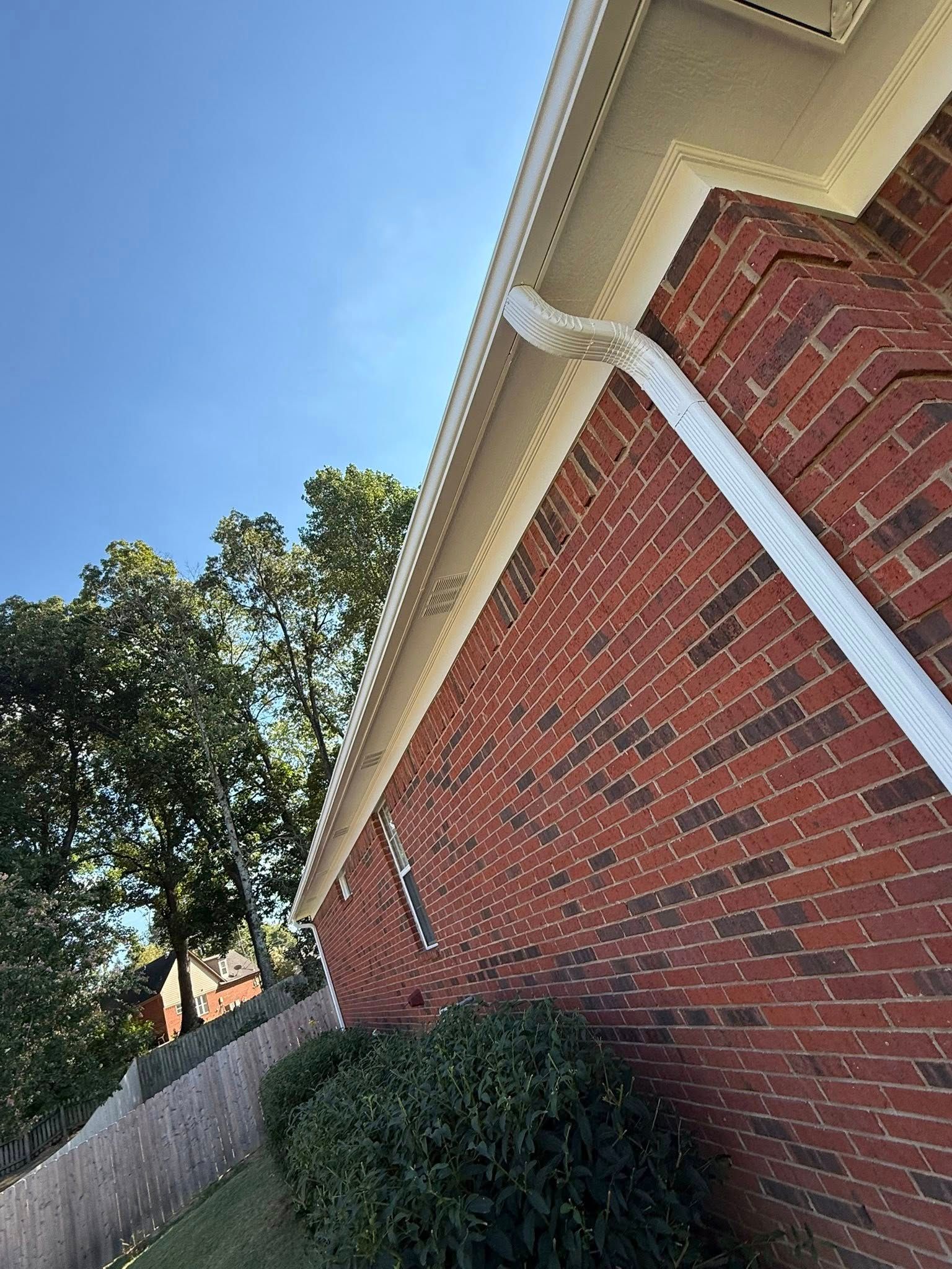 Brick house exterior with white trim, angled roofline, shrub, fence, and blue sky above