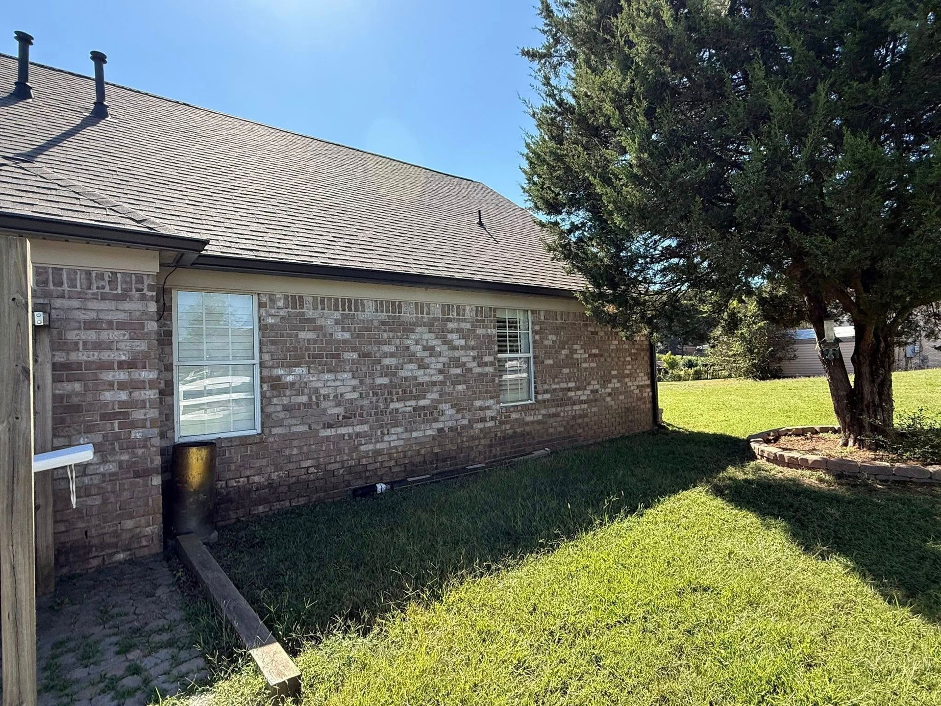 Brick building with a sloped roof, windows, and a grassy yard beside a tree under a blue sky