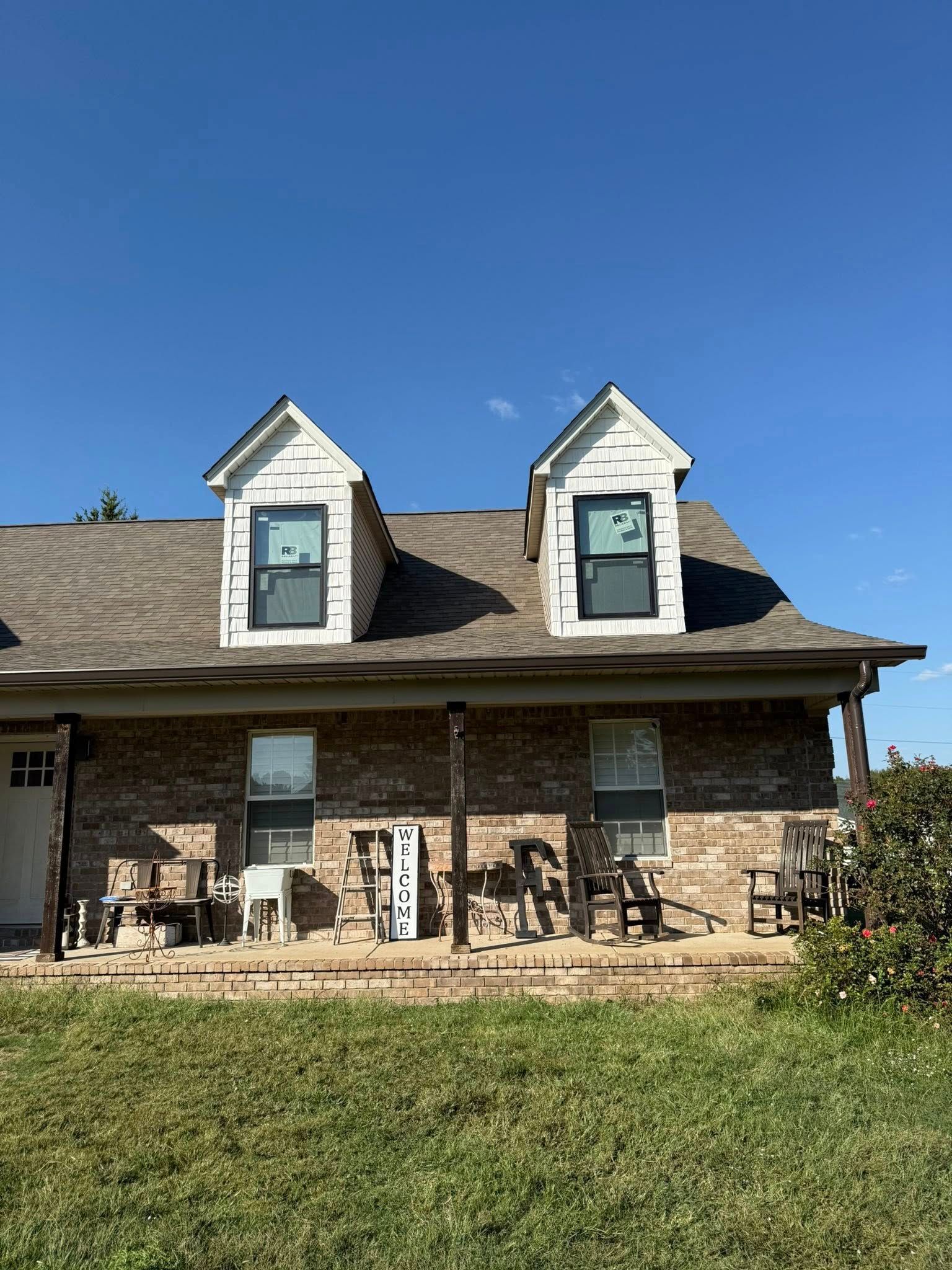 Brick house with dormer windows, porch chairs, and a green lawn under a clear blue sky