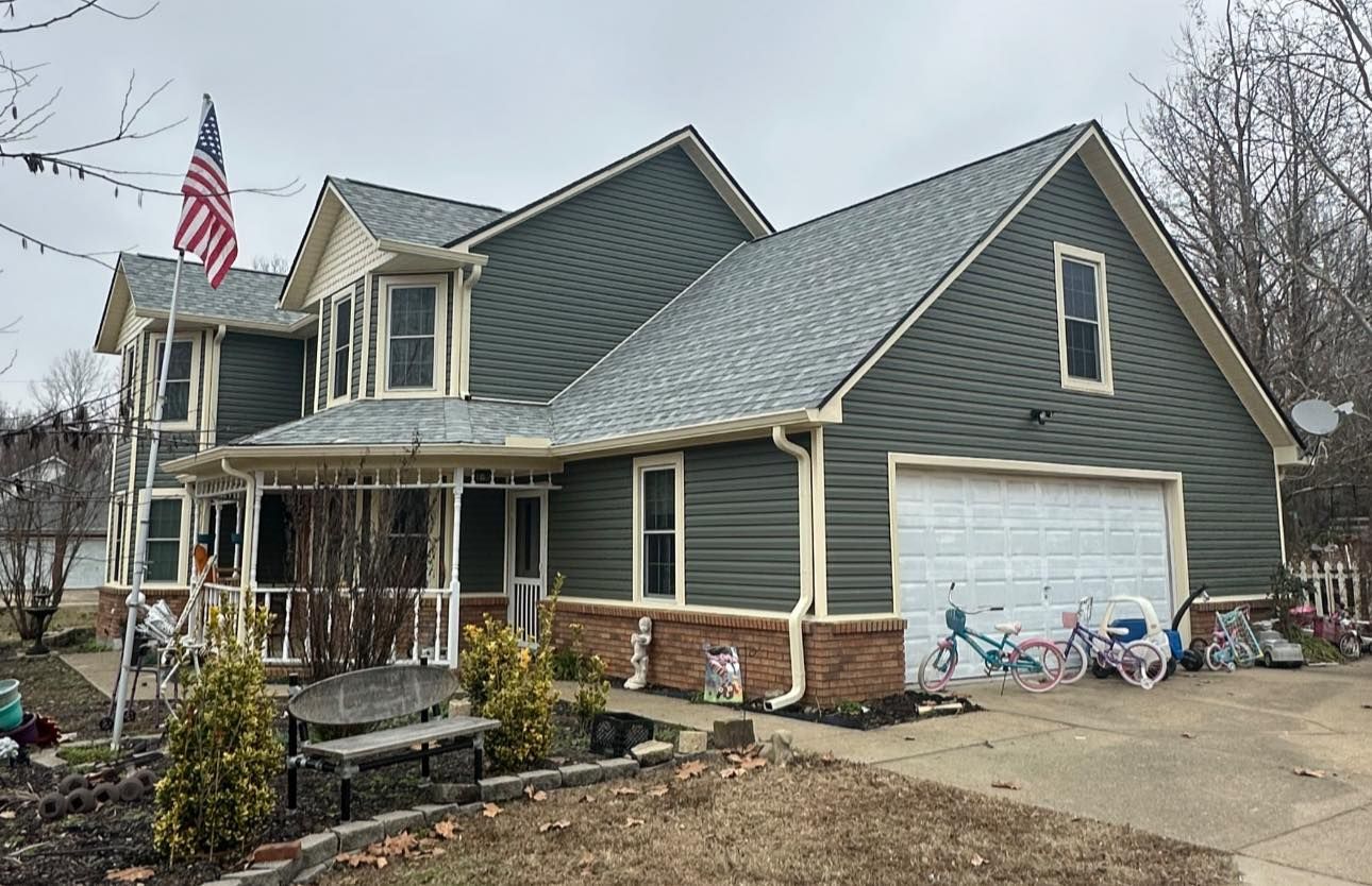 Suburban house with gray siding, front porch, brick accents, and a white garage under a cloudy sky