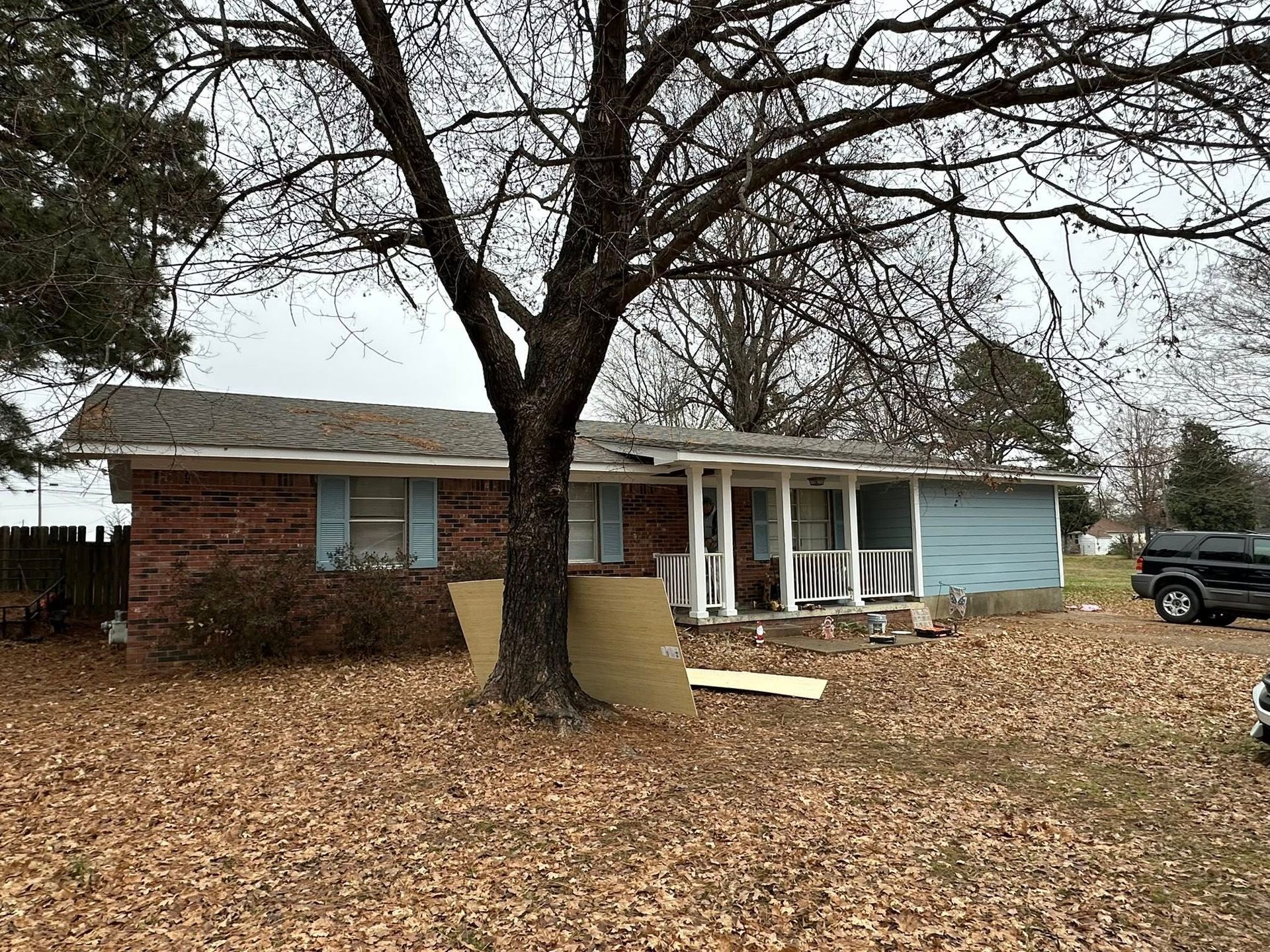 Single-story brick house with light blue siding, large tree in front, and fallen leaves in yard