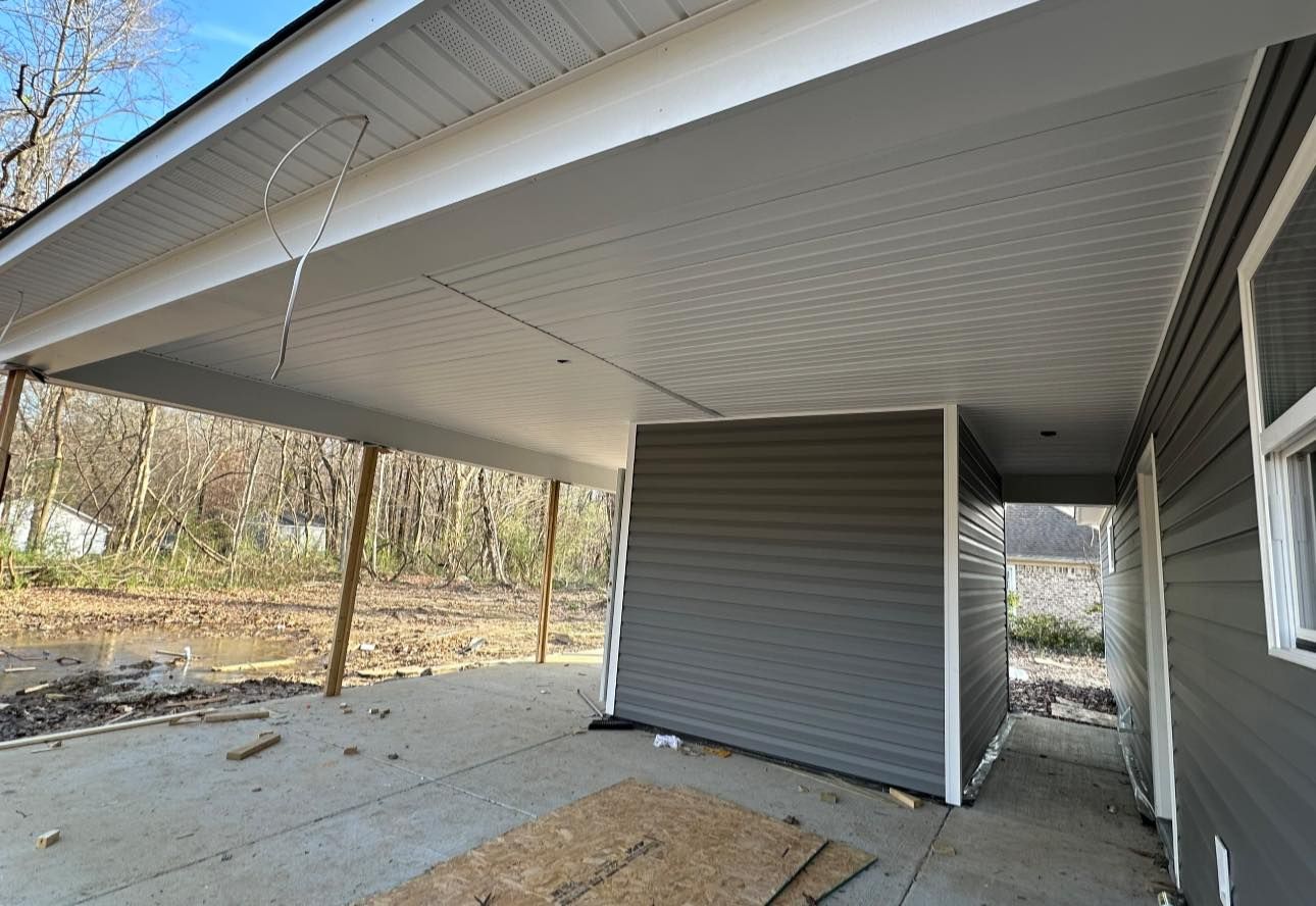 Covered patio under construction beside a house, with gray siding and exposed concrete floor