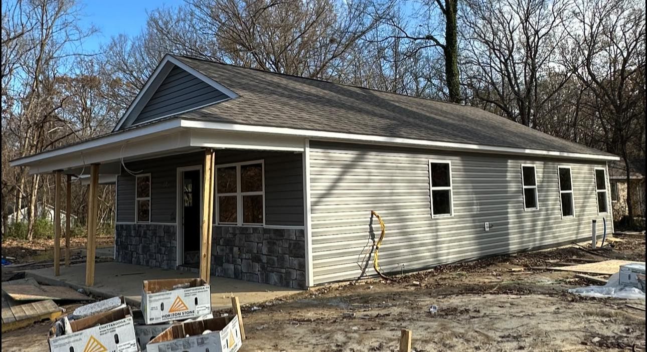 Single-story beige house under construction with stone porch and bare trees around it