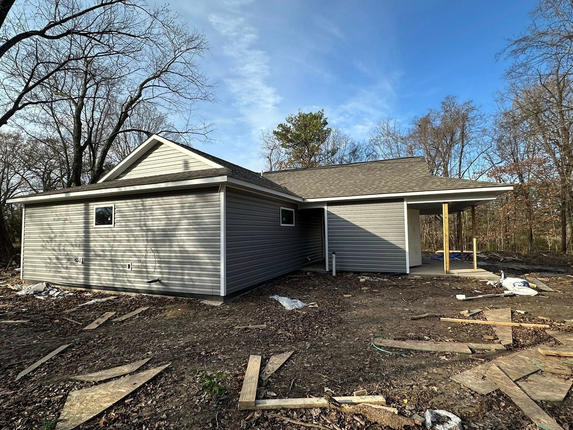 Gray house under construction with unfinished yard and bare trees in the background
