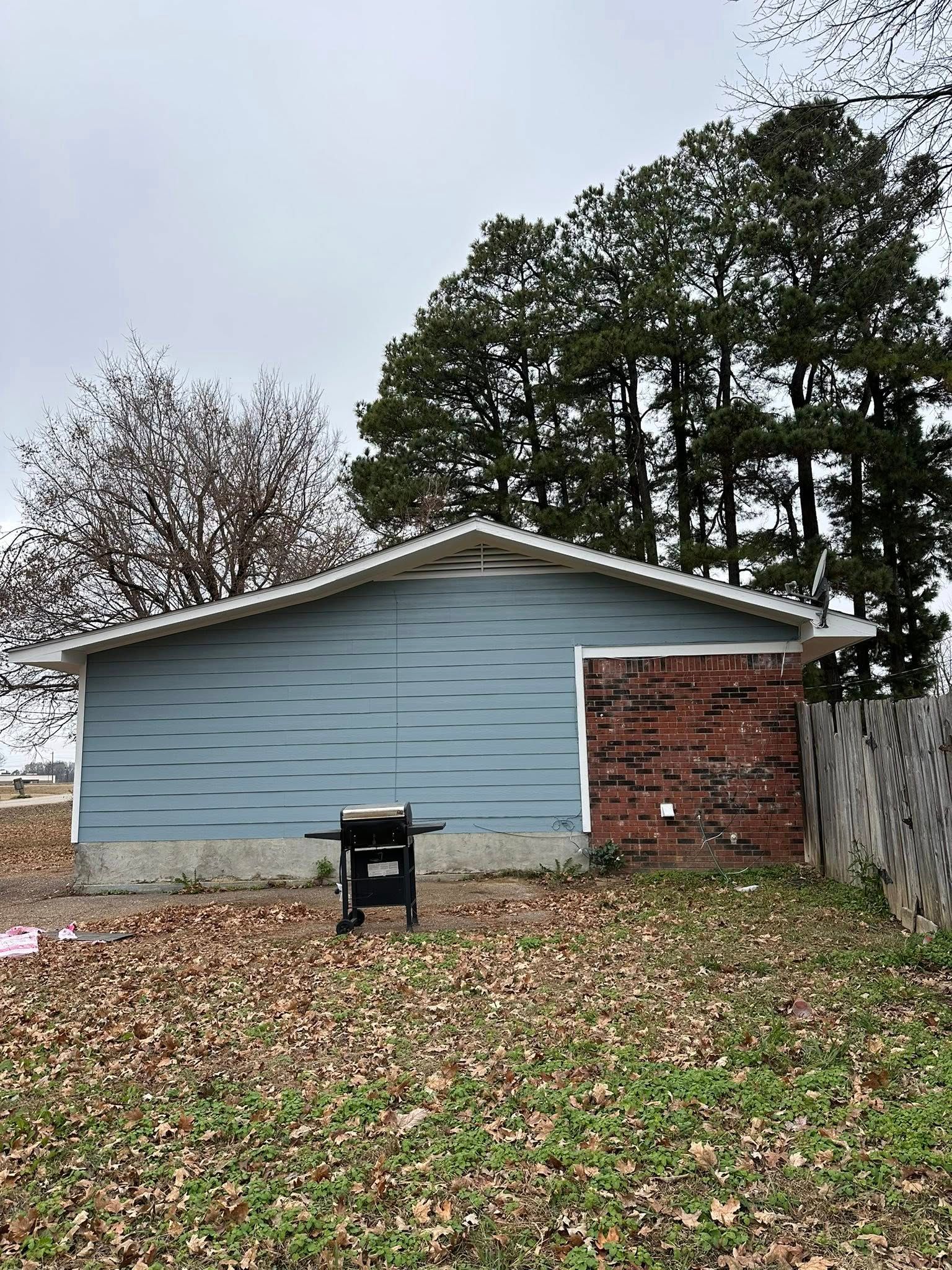 Blue house with brick side wall, backyard grill, and tall trees under an overcast sky