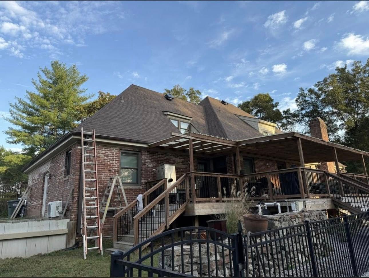 Brick house with dark roof, porches, ladders, and a black fence under a blue sky