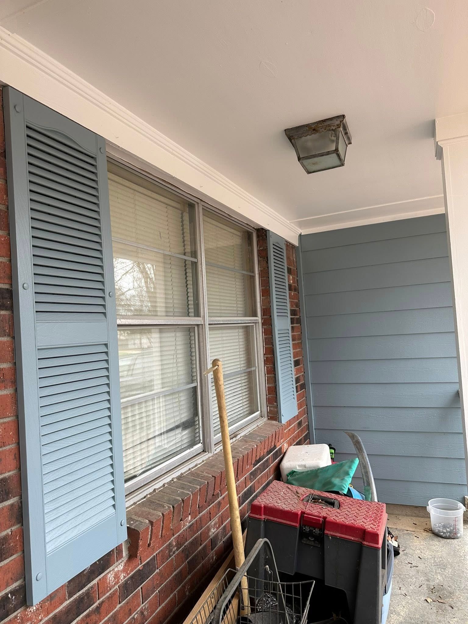 Covered porch with blue siding, brick wall, window shutters, tools, and storage bins.