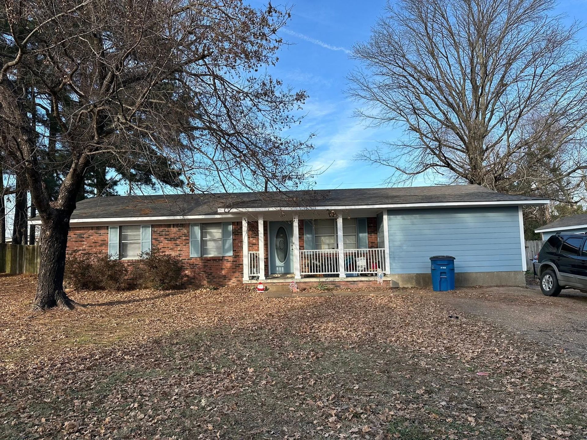 Single-story brick house with a white garage, leaf-covered yard, and blue recycling bin under a blue sky