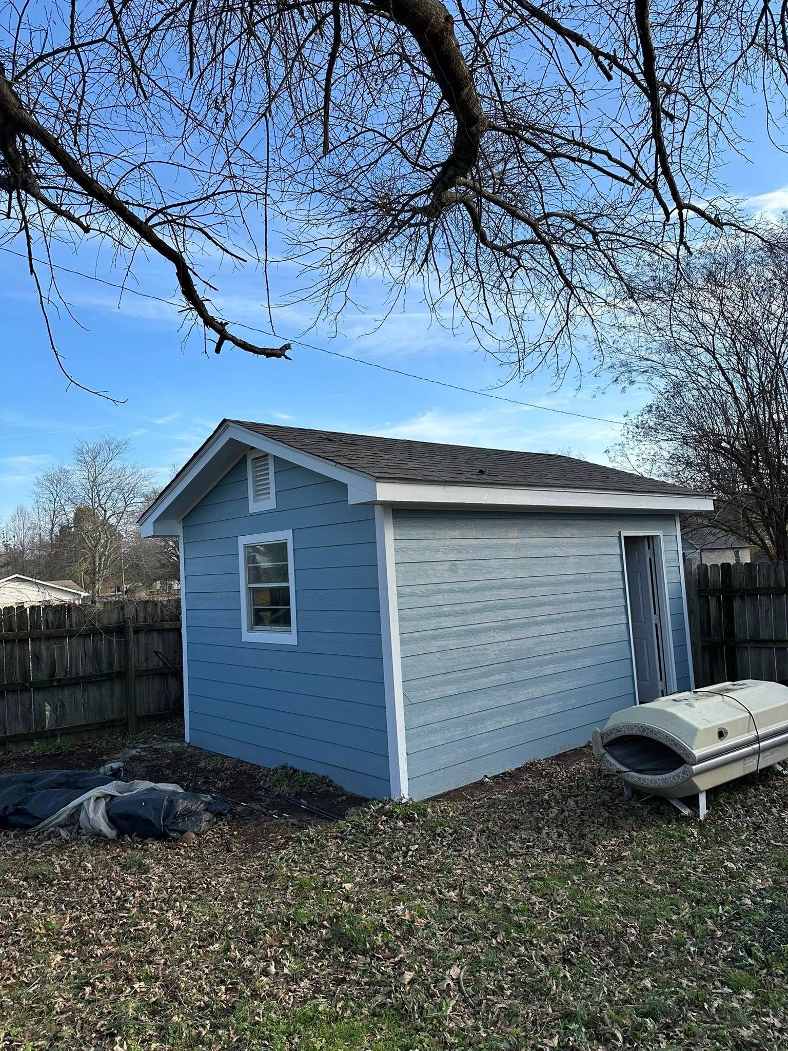 Small blue and white shed in a backyard under leafless tree branches, with a storage box nearby.