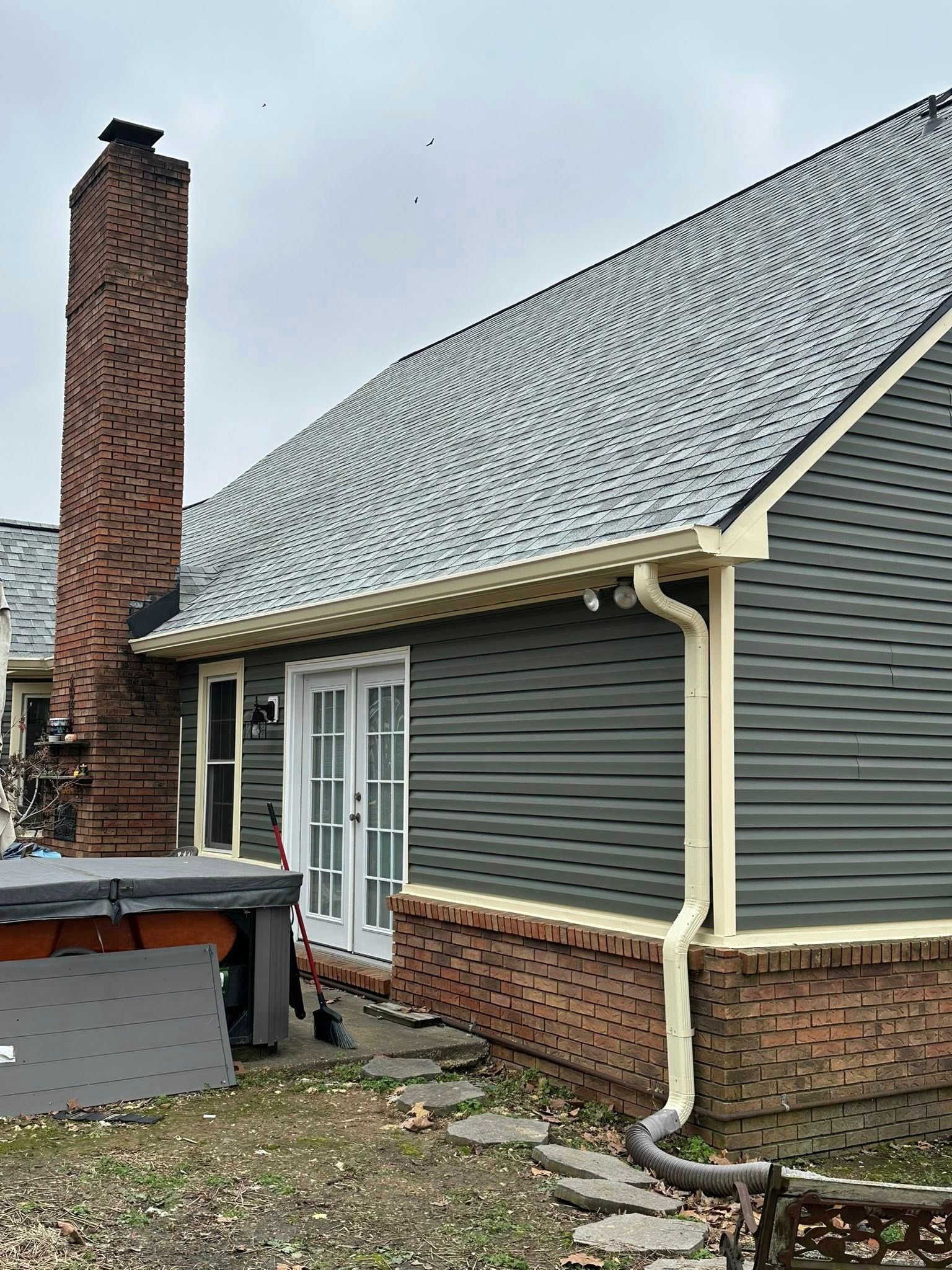 Backyard view of a house with gray siding, brick chimney, and a shingled roof after rain.