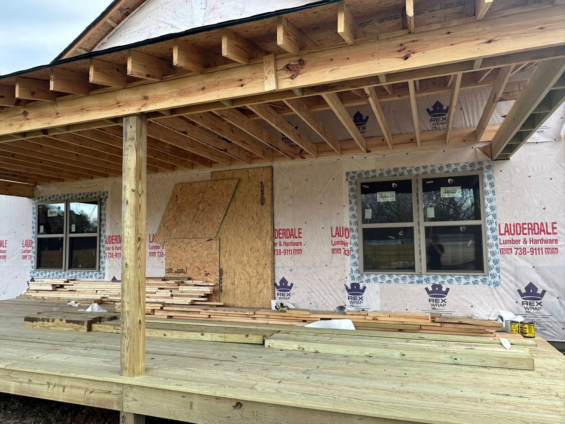 House under construction with exposed framing, plywood, and wrapped exterior walls on a covered porch