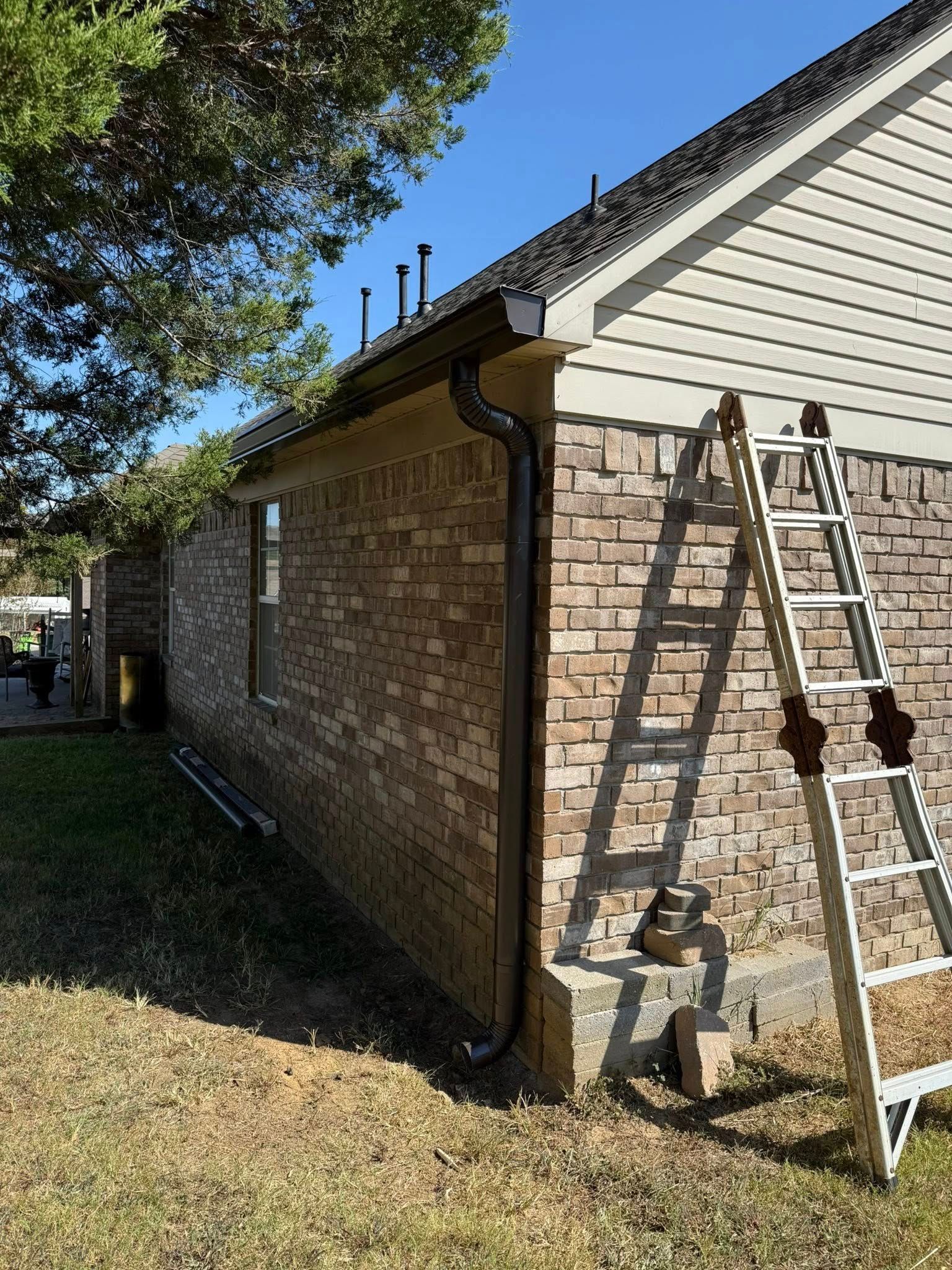 Brick house side wall with ladder leaning against it, gutter downspout, and roof edge in a yard.