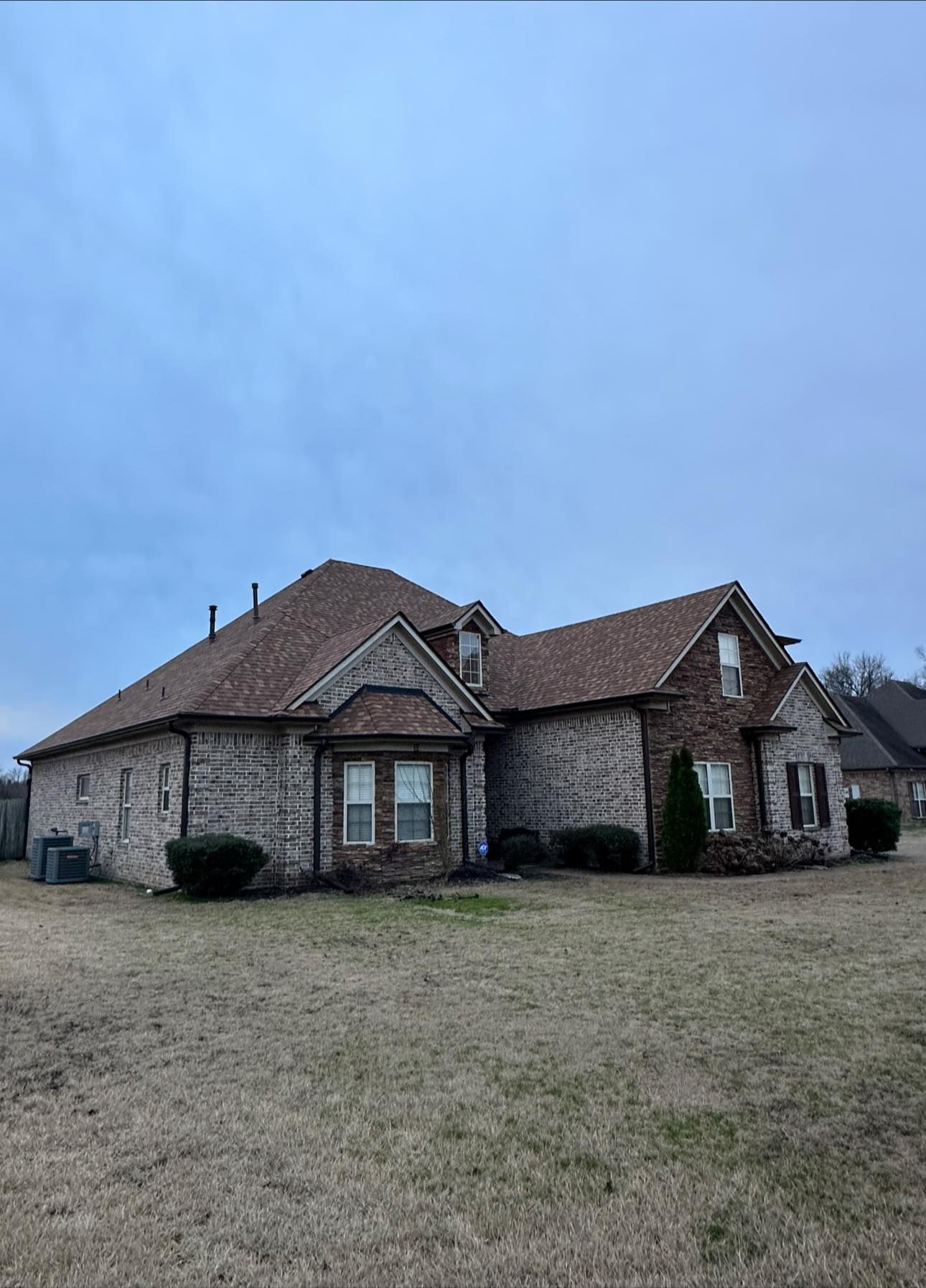 Single-story brick house with a dark shingled roof and large yard under a cloudy blue sky