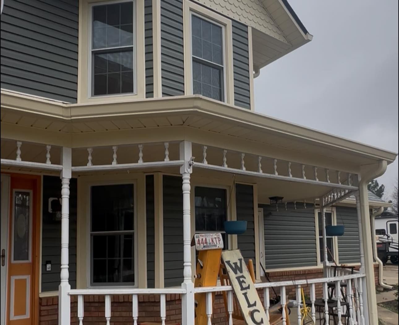 Porch of a gray Victorian house with white trim and a hanging welcome sign.