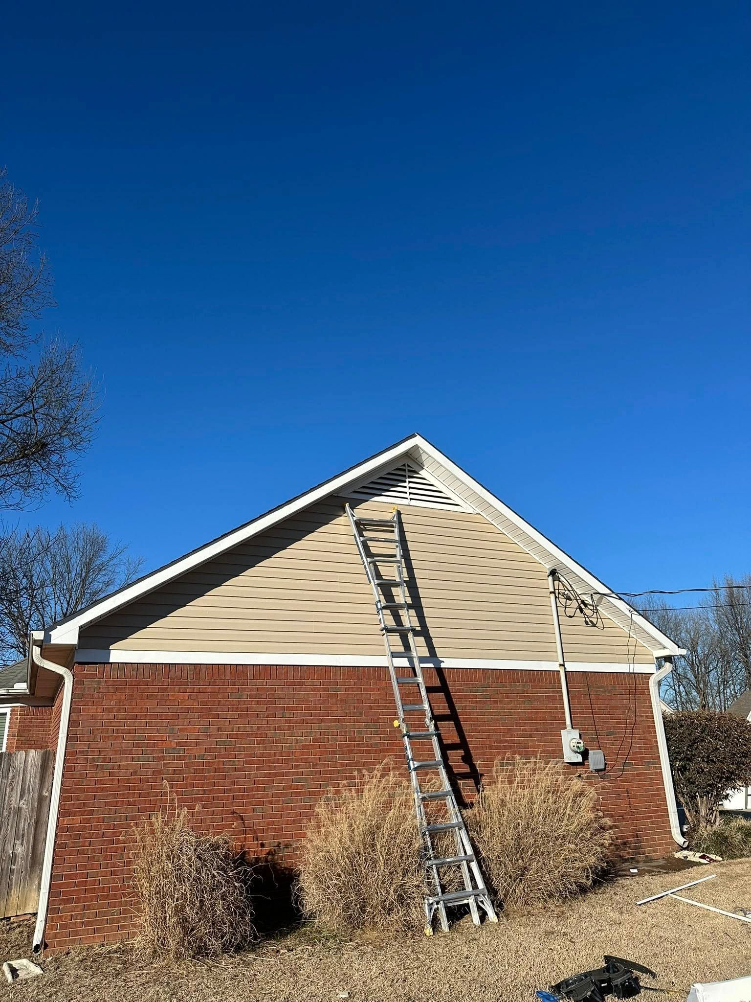 Brick house with tan siding and a ladder leaning against the front gable under a clear blue sky