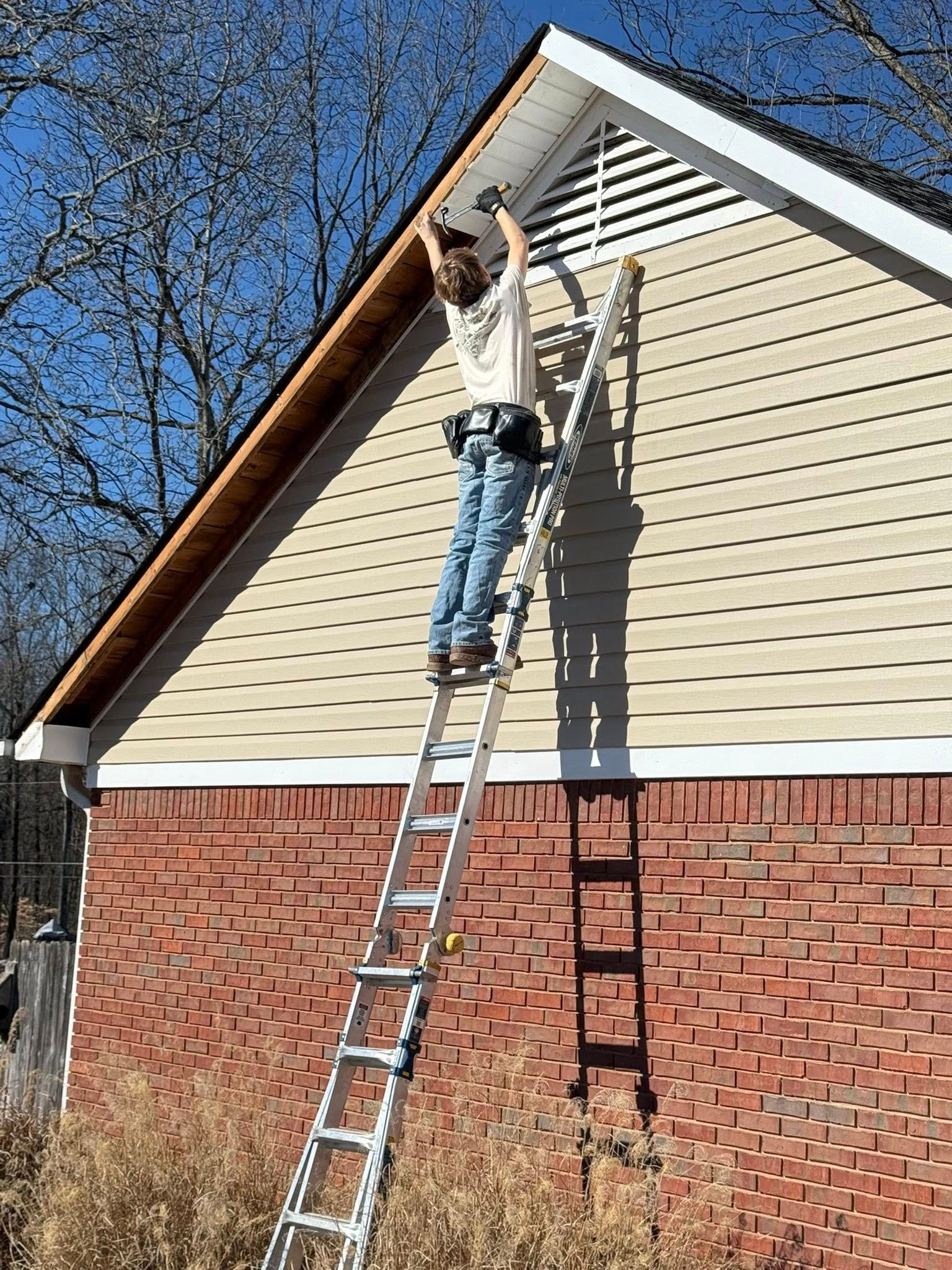 Person on a ladder repairing siding on a house exterior under a gable roof.