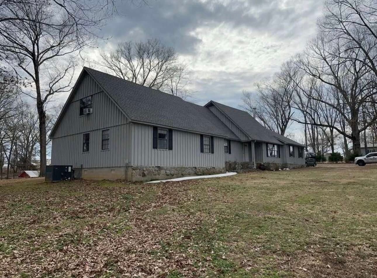 Gray house with a steep roof on a grassy lot under a cloudy sky, surrounded by leafless trees
