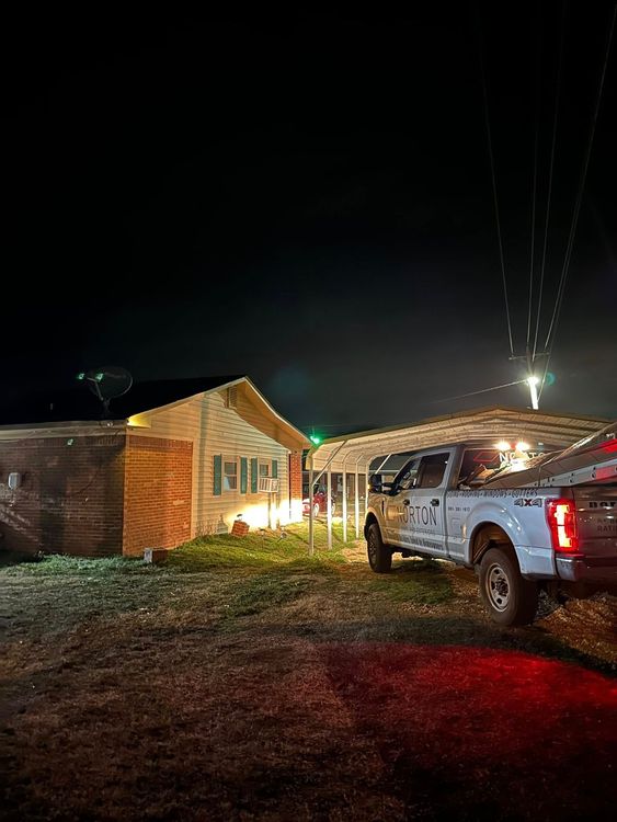 White pickup truck beside a lit brick house at night, with red taillights glowing on muddy ground