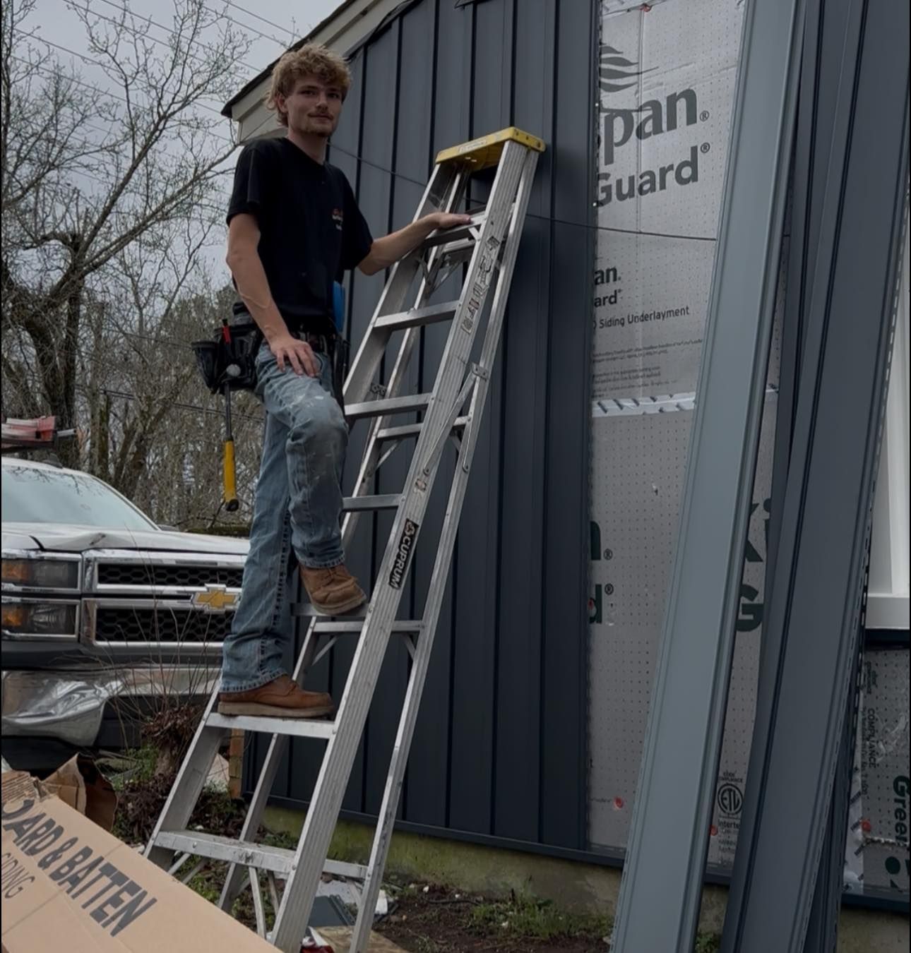 Worker stands on a ladder installing gray siding on a building exterior.