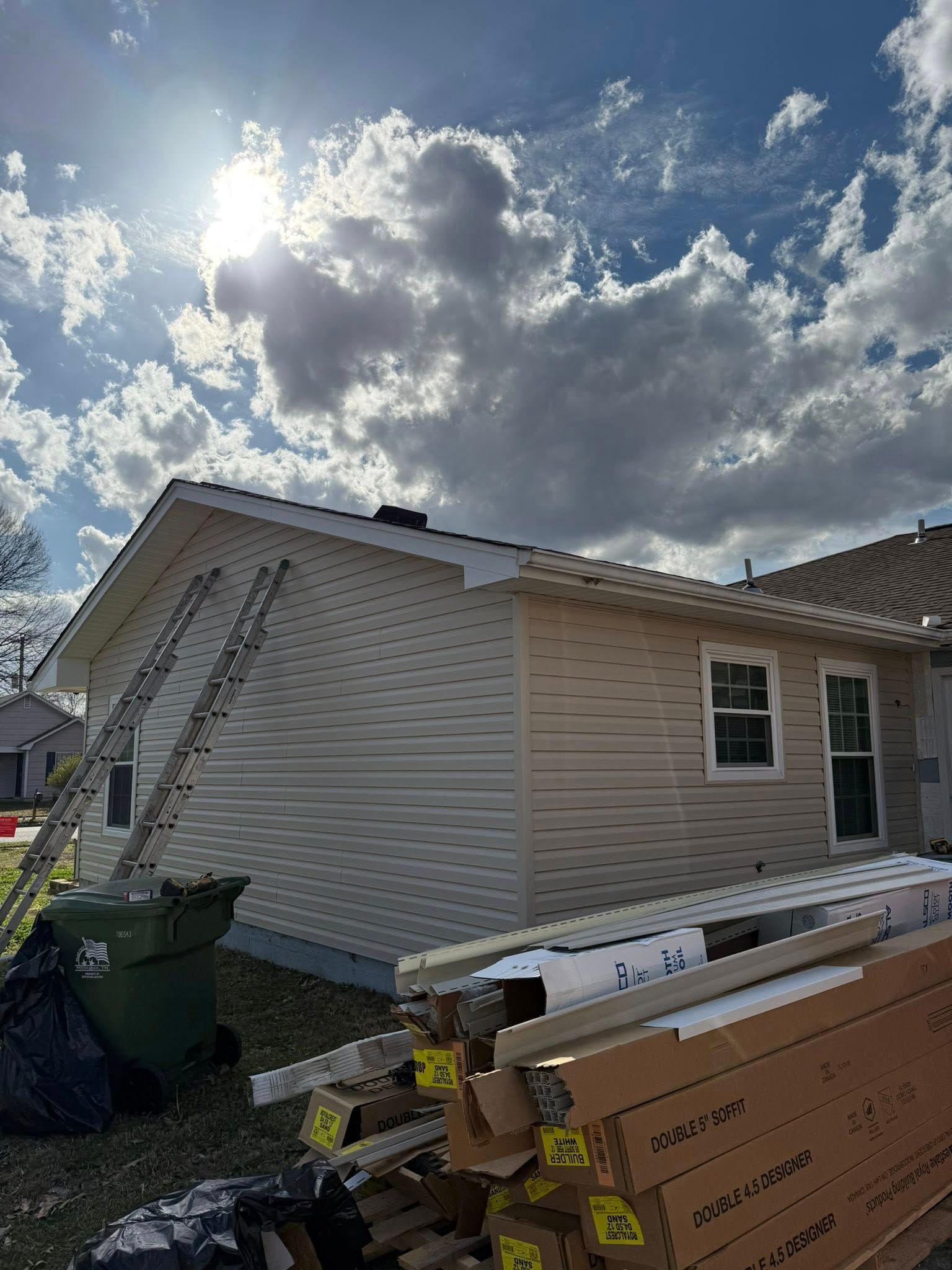 House exterior with white siding, ladders, and construction materials under a bright cloudy sky