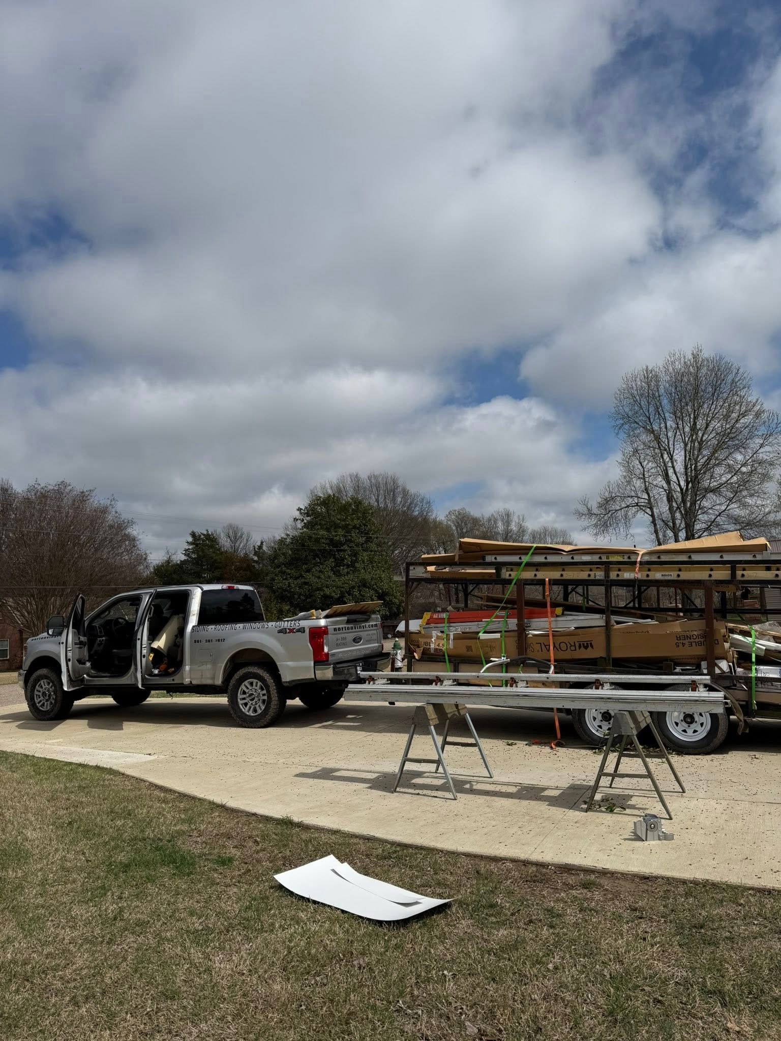 White pickup truck with open doors beside a utility trailer carrying wooden framing at a work site