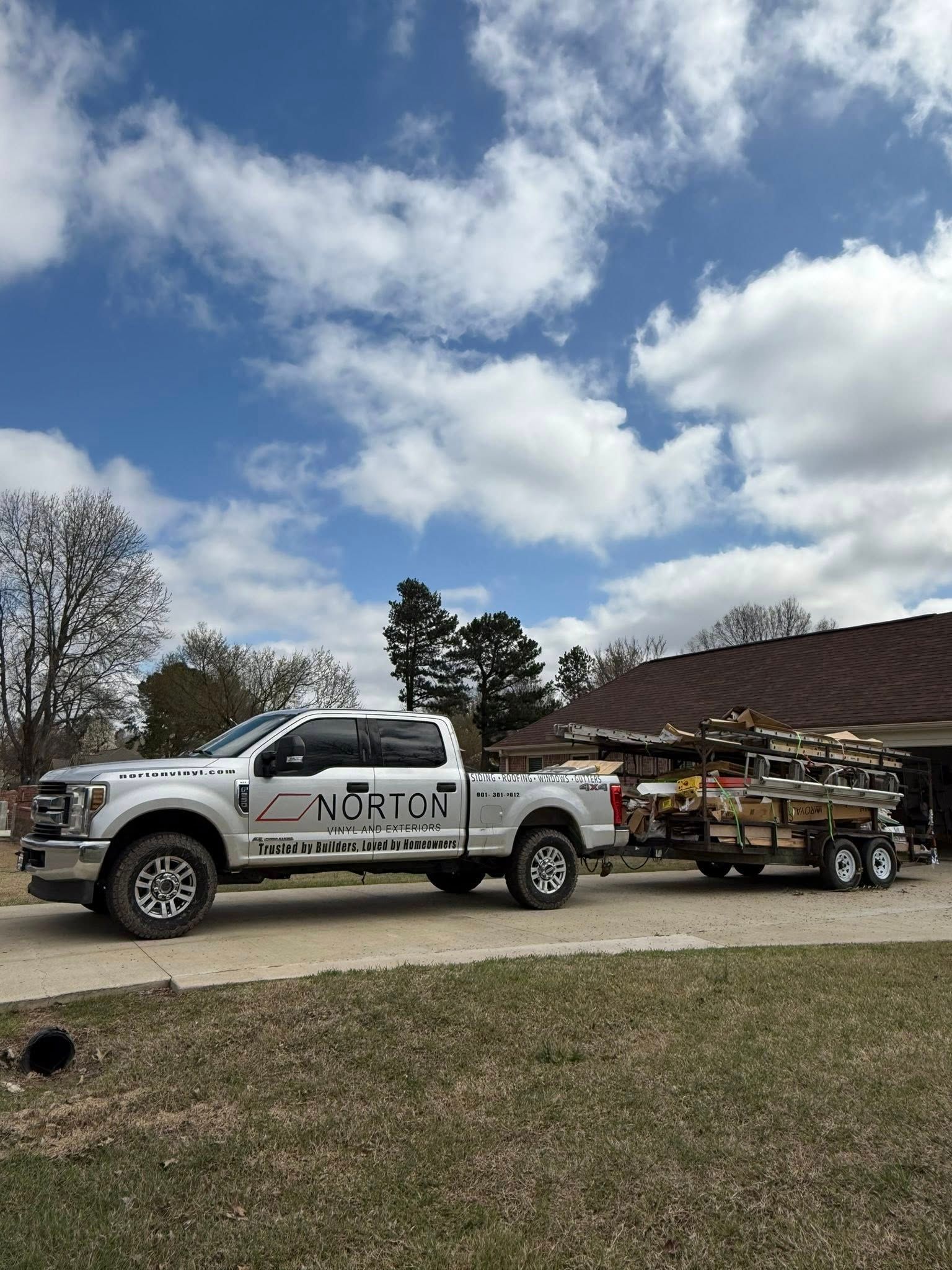 White pickup truck towing a loaded trailer parked beside a house under a cloudy sky