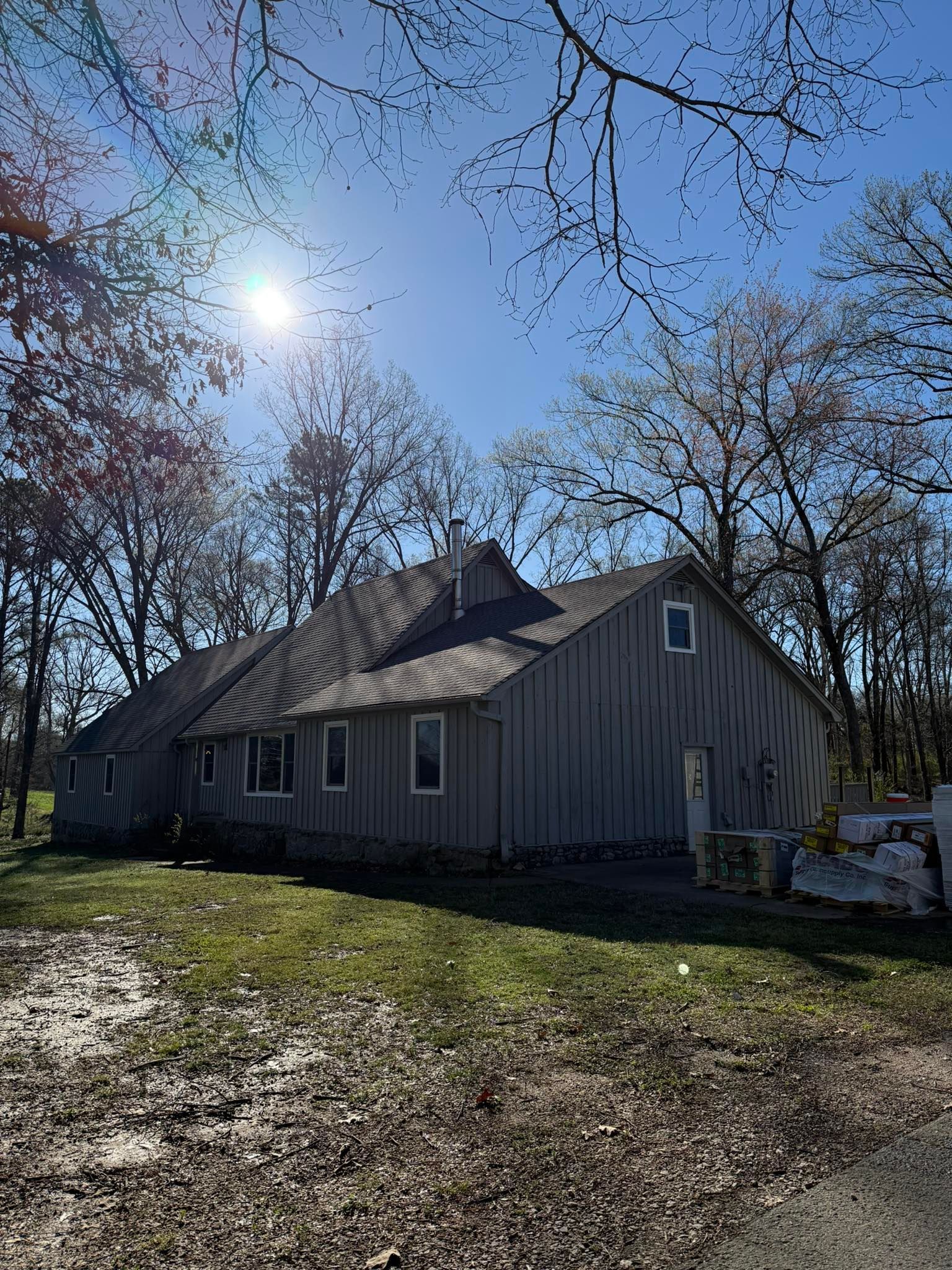 Gray house with a steep roof in a sunny wooded yard, with bare trees and blue sky.